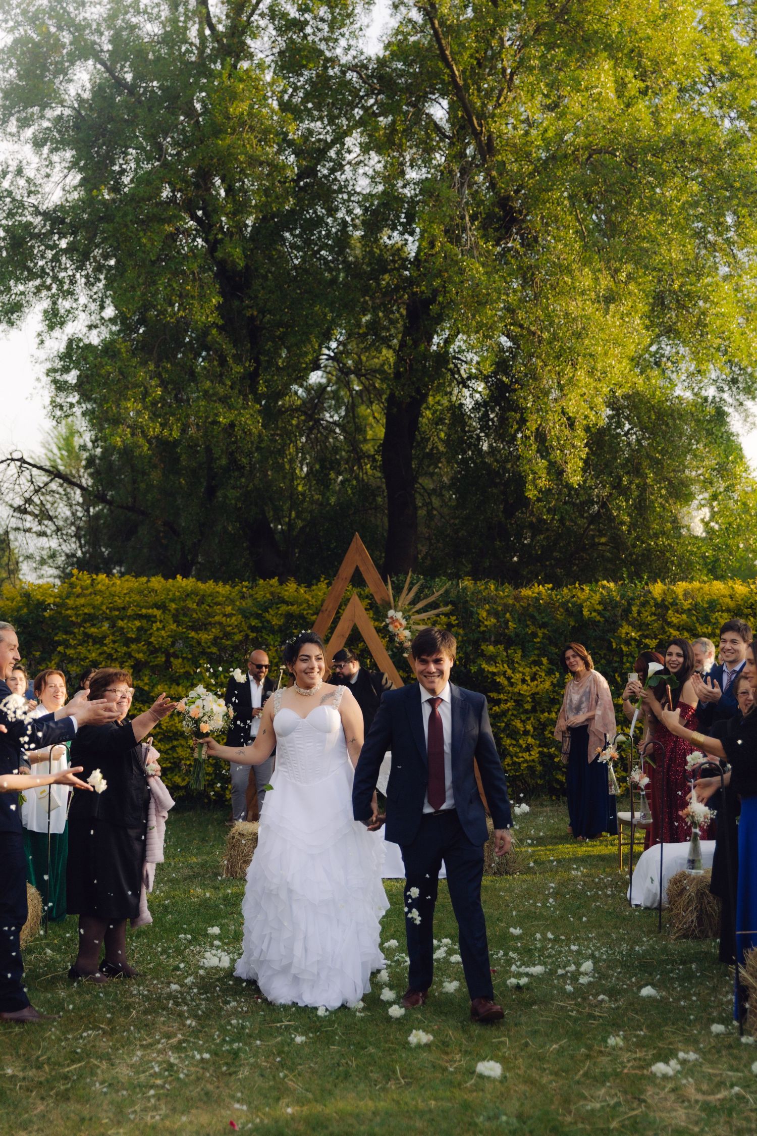 Happy newlyweds hold hands during joyful confetti recessional at outdoor bohemian garden wedding.
