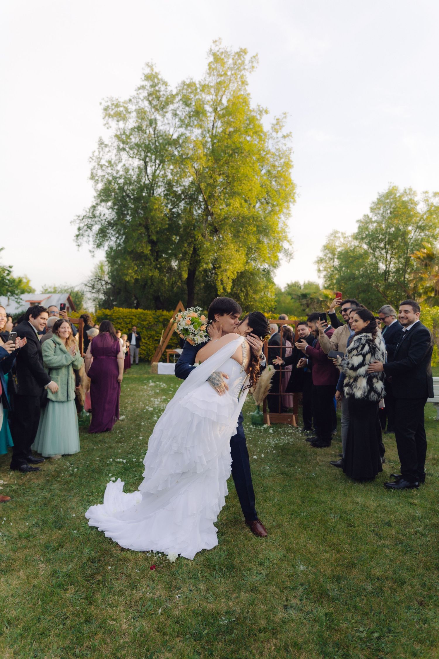 Groom dips bride for a romantic kiss after outdoor wedding ceremony surrounded by cheering guests.