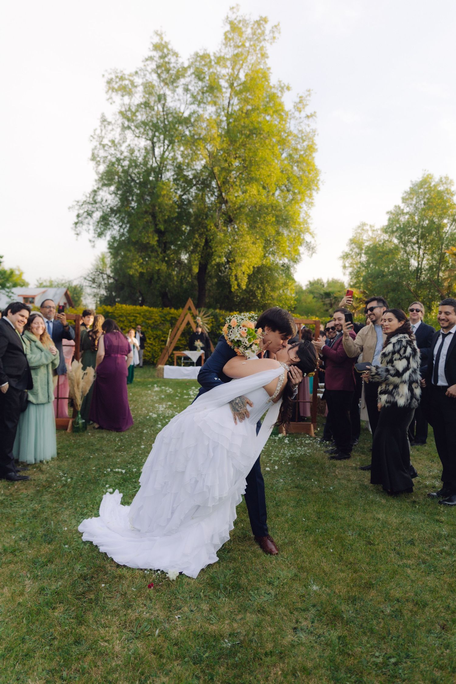 Newlyweds share a dip kiss on green lawn after wedding ceremony with guests watching in background.