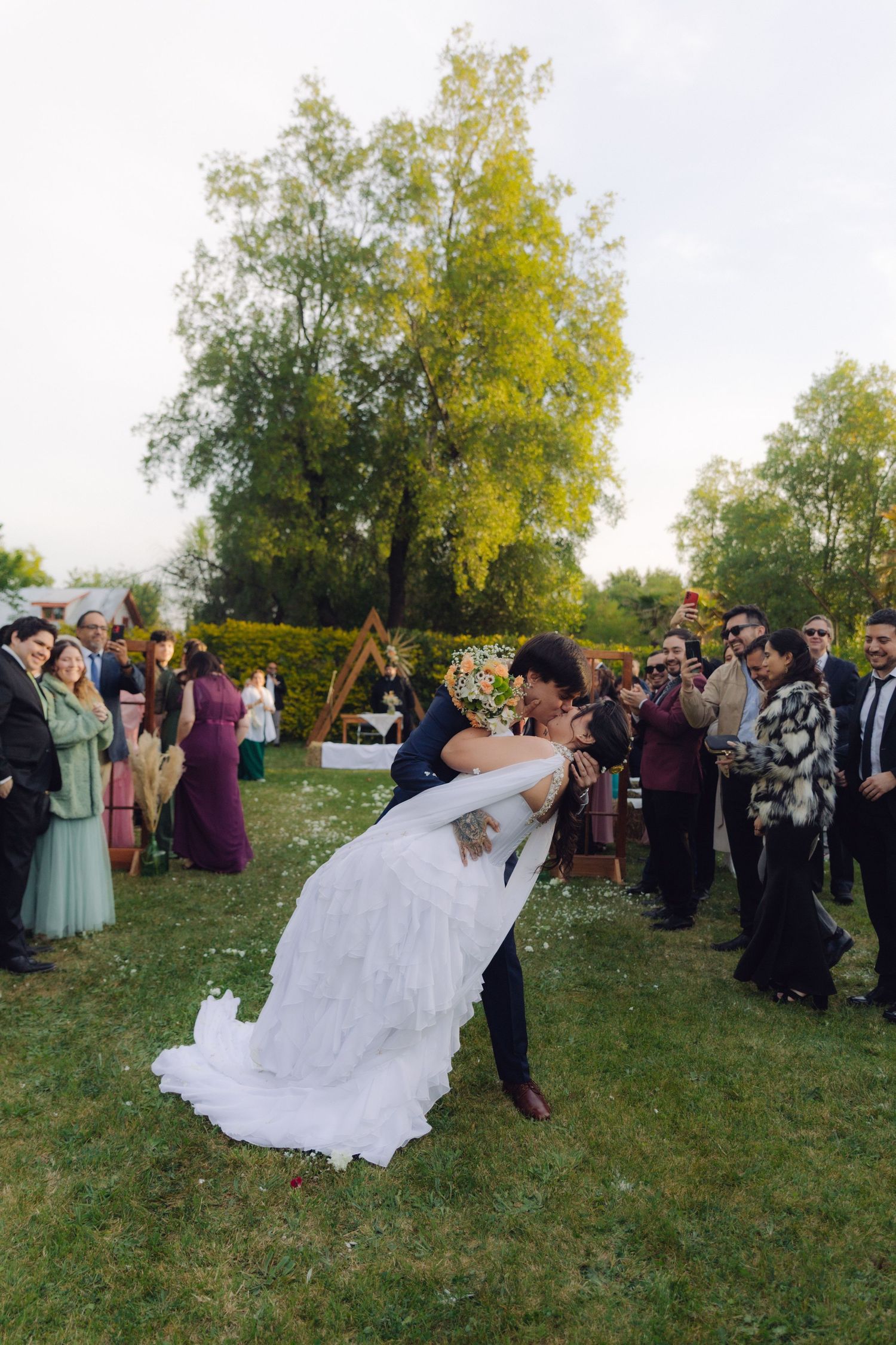Groom dramatically dips bride for a kiss outdoors after wedding ceremony with guests applauding around them.