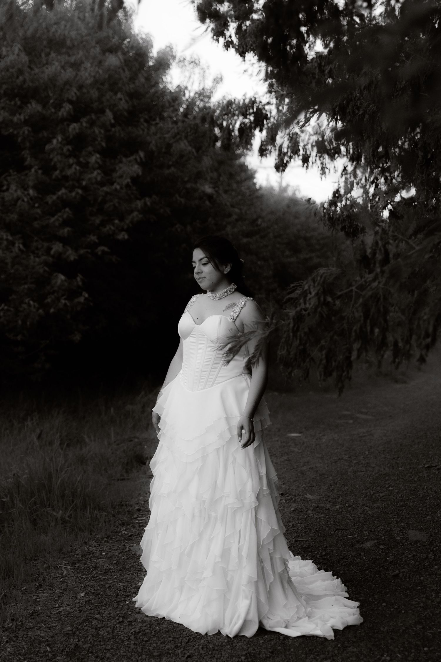 Elegant bride in white ruffled gown poses in black and white portrait among dark forest trees.