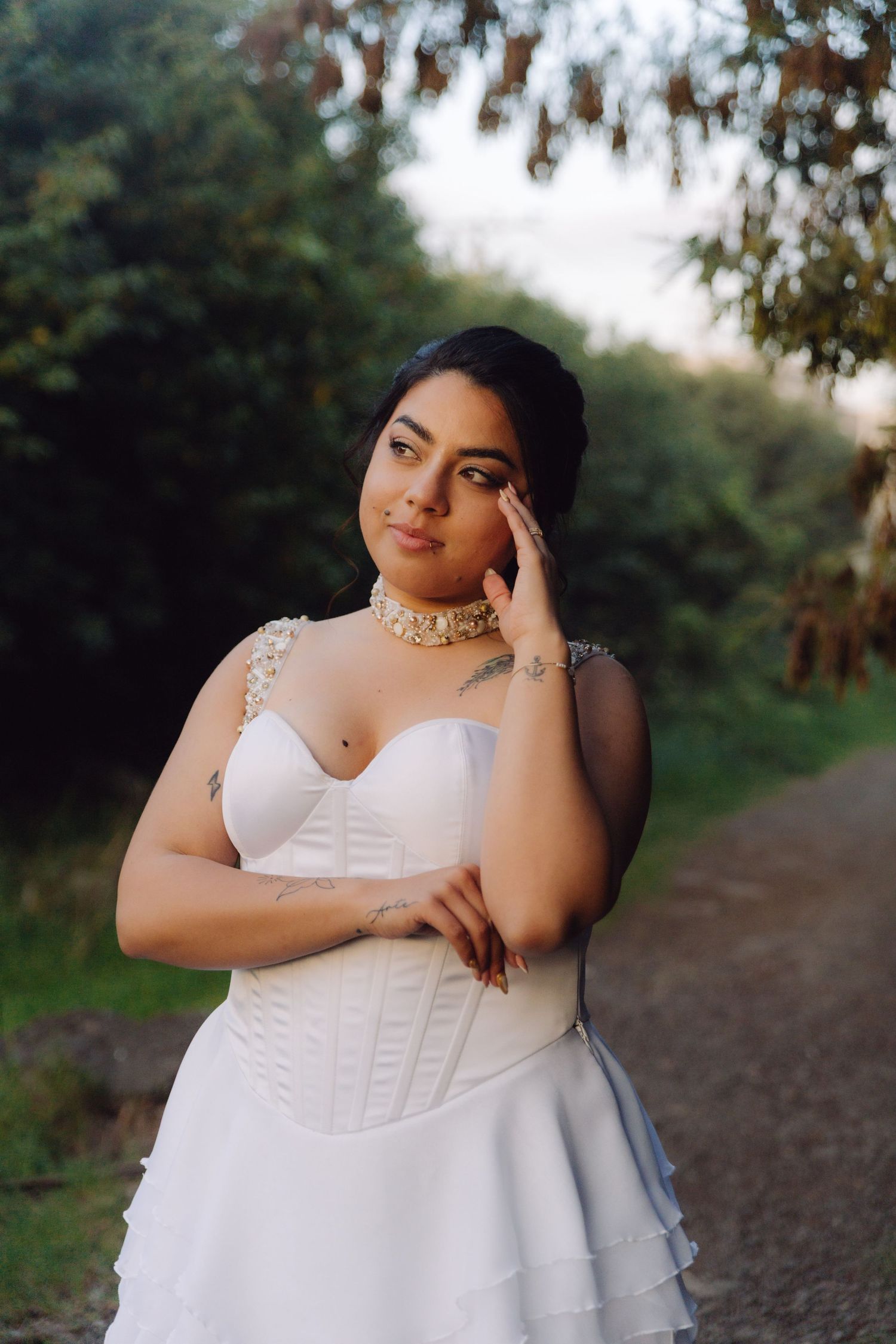 Beautiful tattooed bride in white corset wedding gown with pearl choker poses along a garden path.