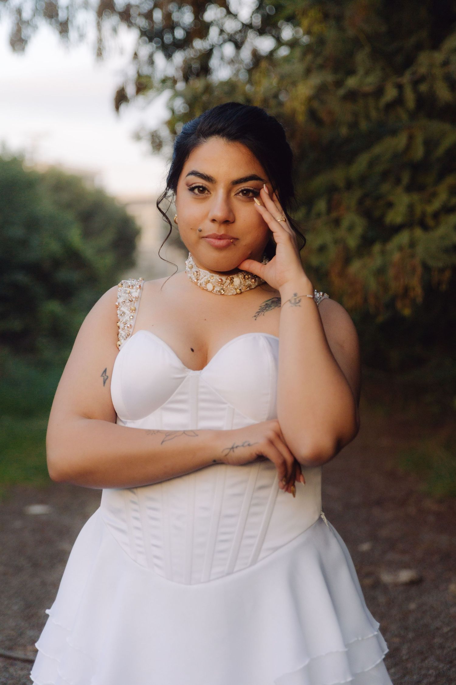 Stunning bride in white corset gown with pearl choker necklace poses gracefully along a tree-lined path.