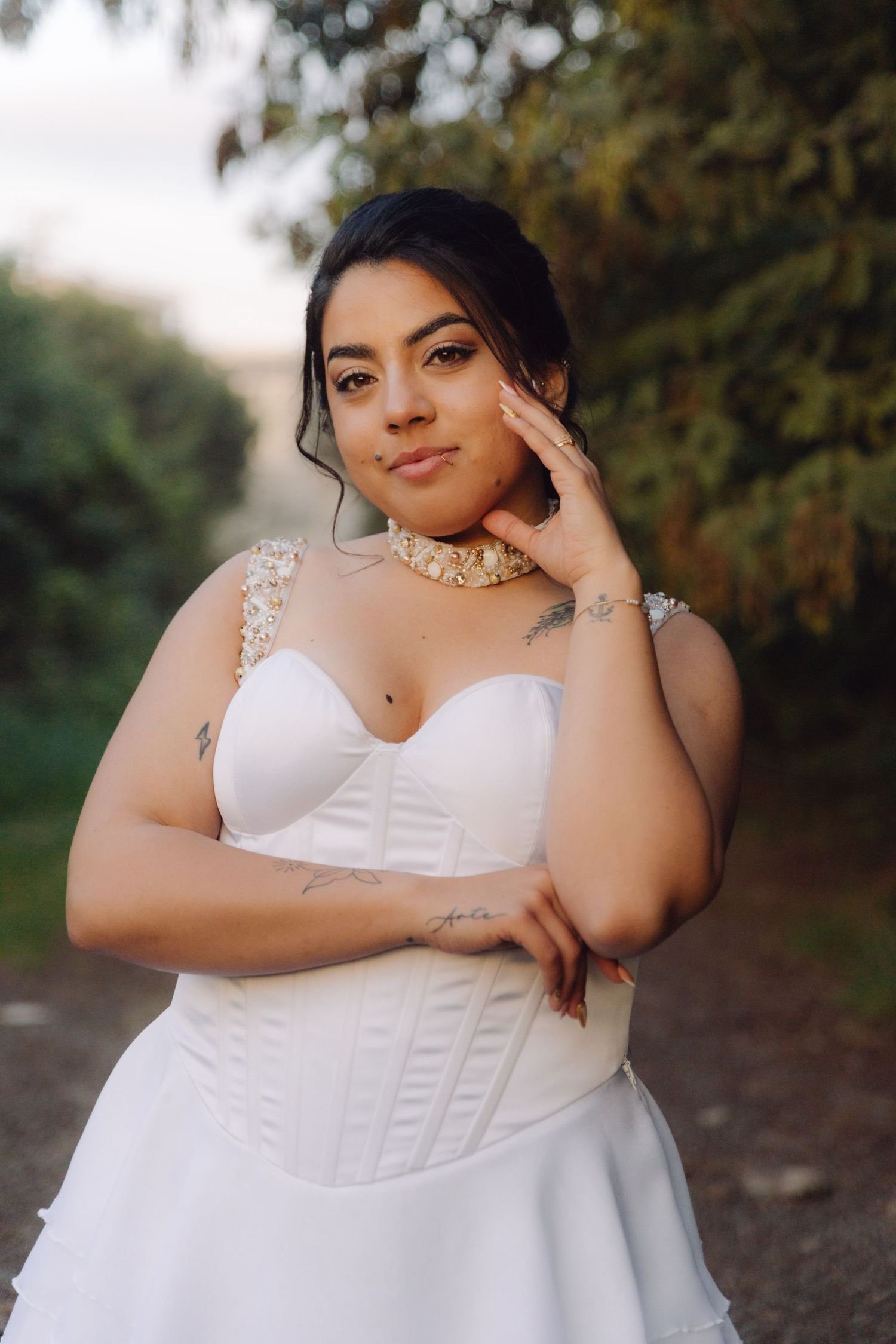 Tattooed bride in white corset wedding dress and pearl choker poses confidently on an outdoor garden path.