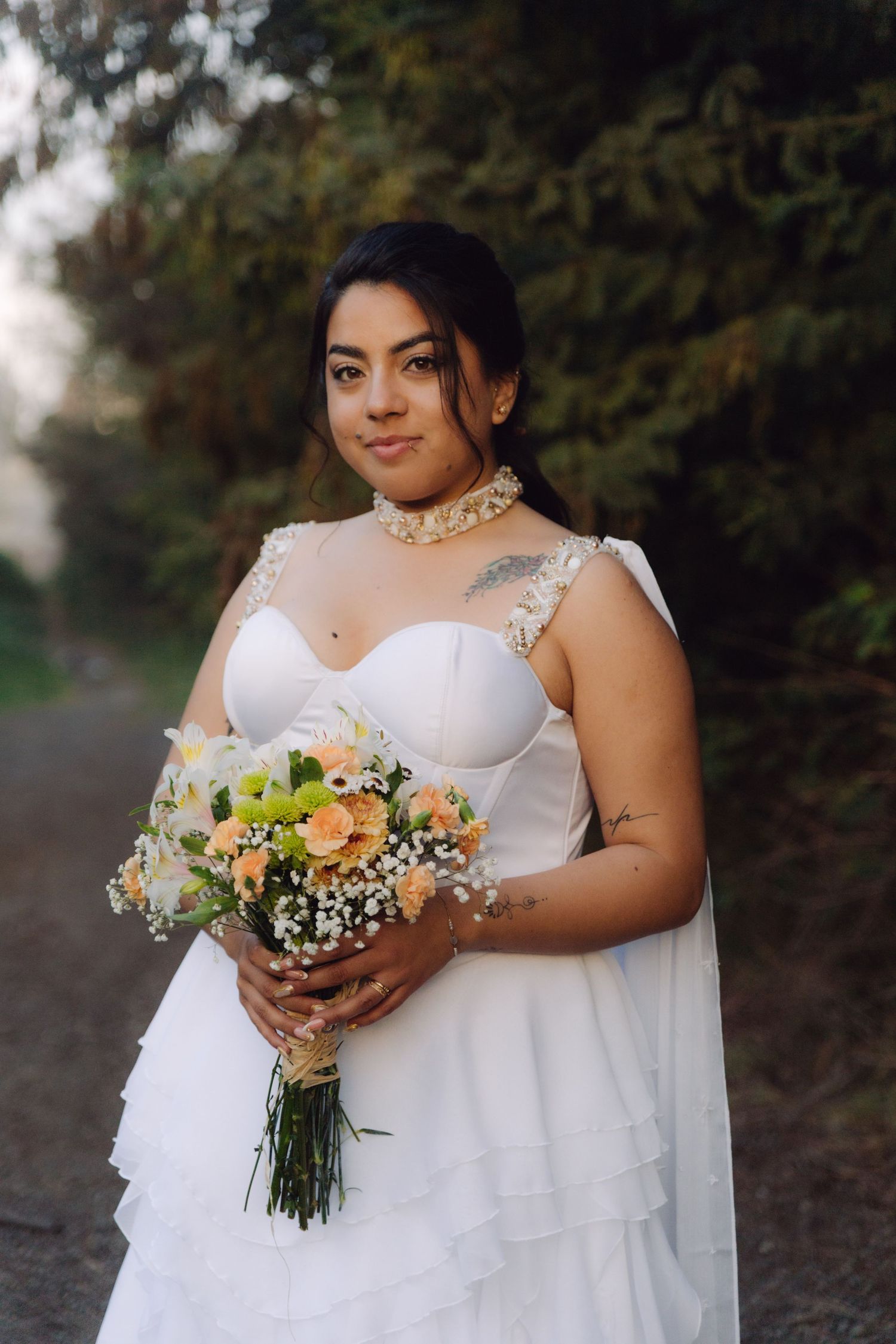 Bride in white ruffled gown holds peach and white bouquet while posing outdoors at golden hour.