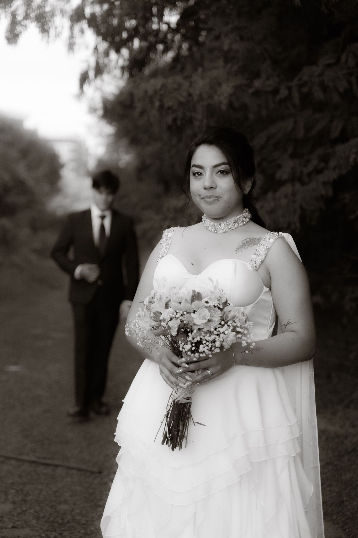 Black and white photo of bride holding bouquet with groom walking in background along a tree-lined path.