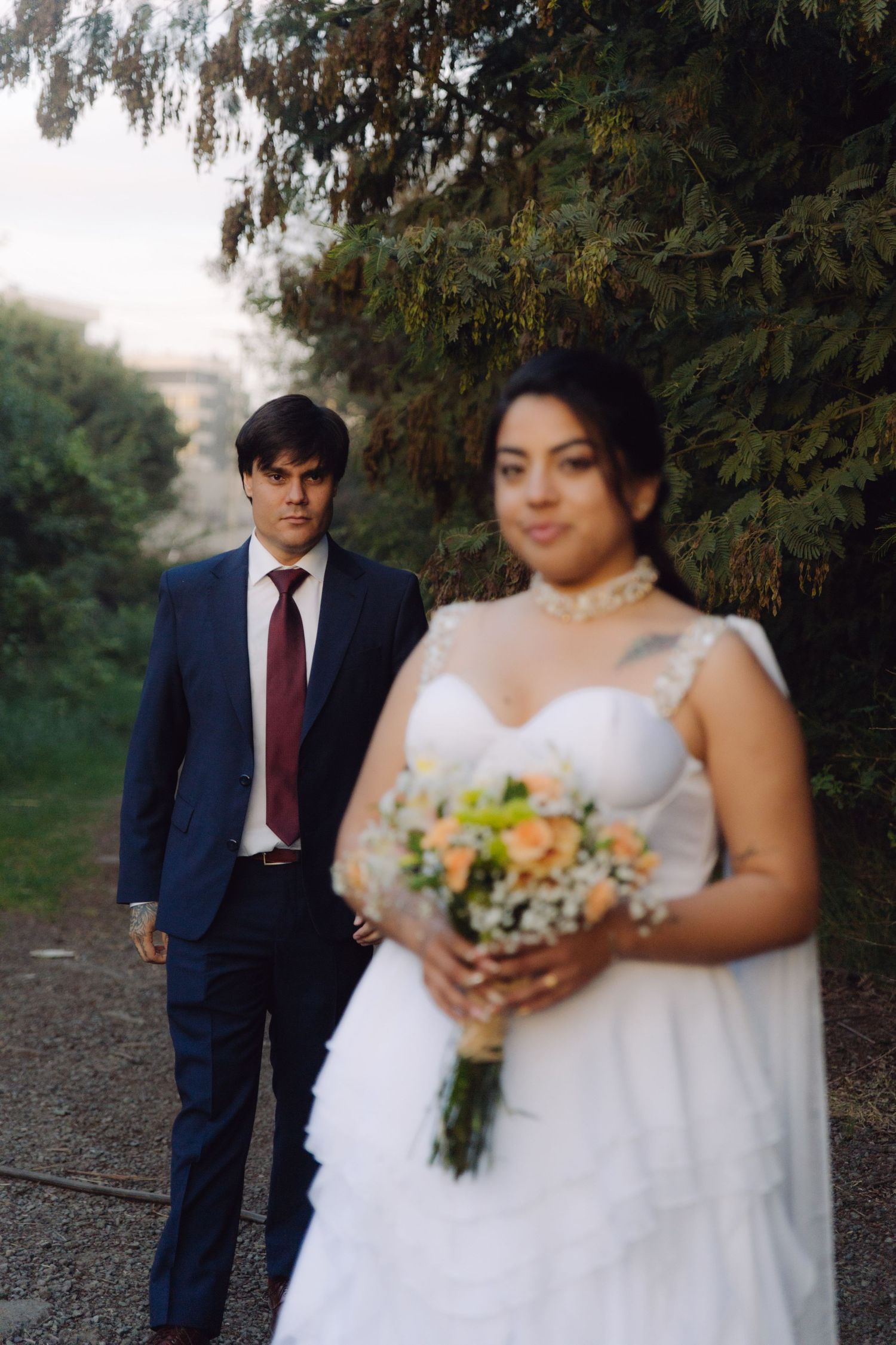 Groom in navy suit walks toward bride holding bouquet in foreground along a scenic tree-lined path.