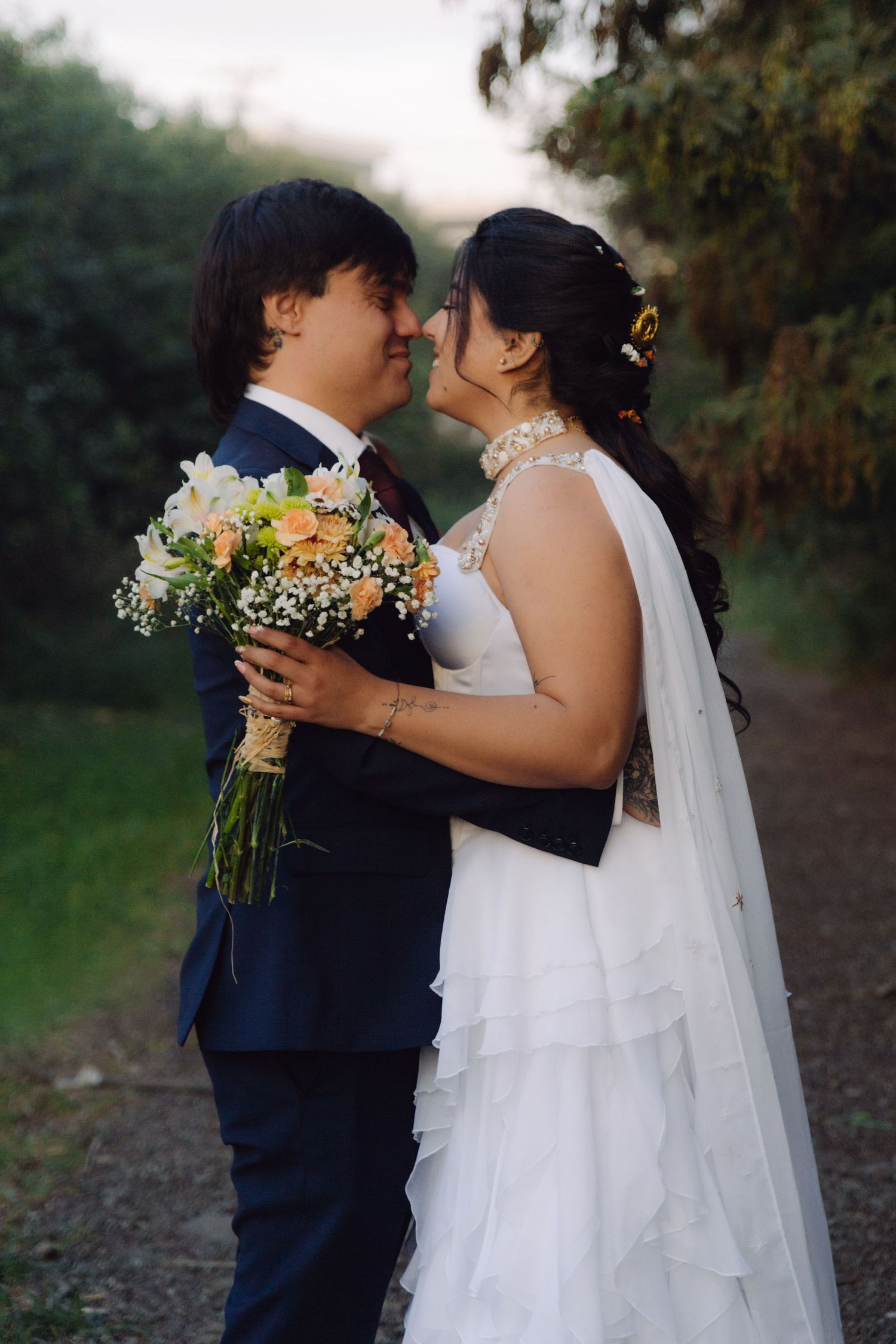 Bride and groom share a romantic almost-kiss while holding a floral bouquet in a lush garden setting.