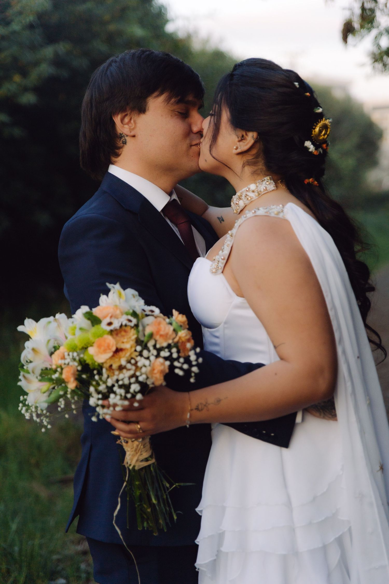 Bride and groom share a kiss while holding a peach and white floral bouquet in an outdoor garden.