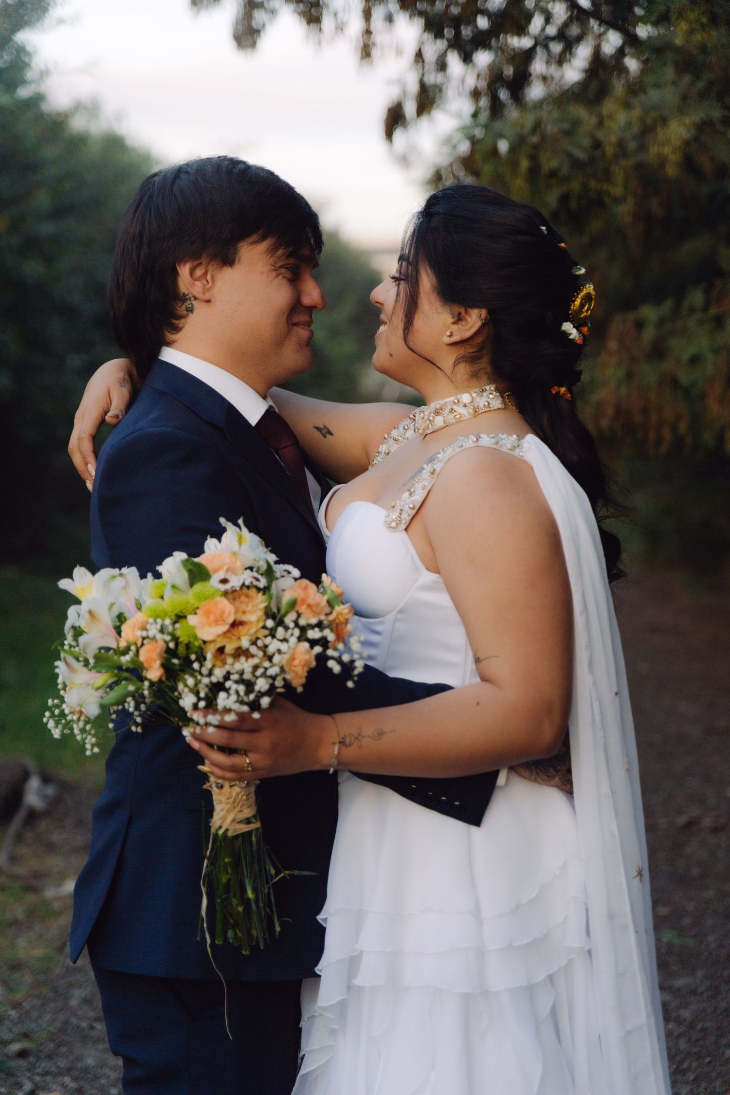 Smiling bride and groom embrace while holding a bouquet on a tree-lined path at sunset.