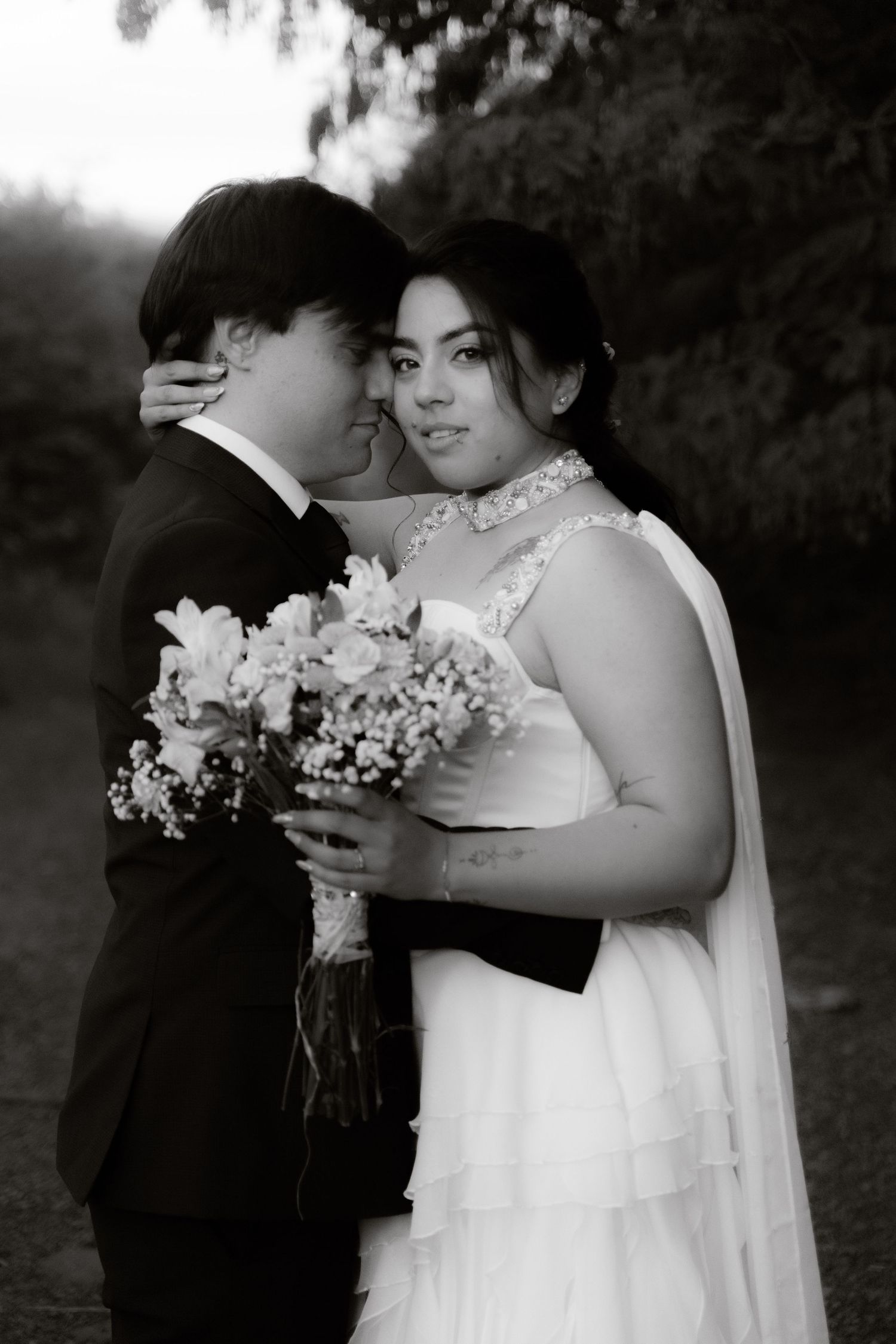 Black and white photo of a bride and groom embracing tenderly, holding a floral bouquet outdoors.