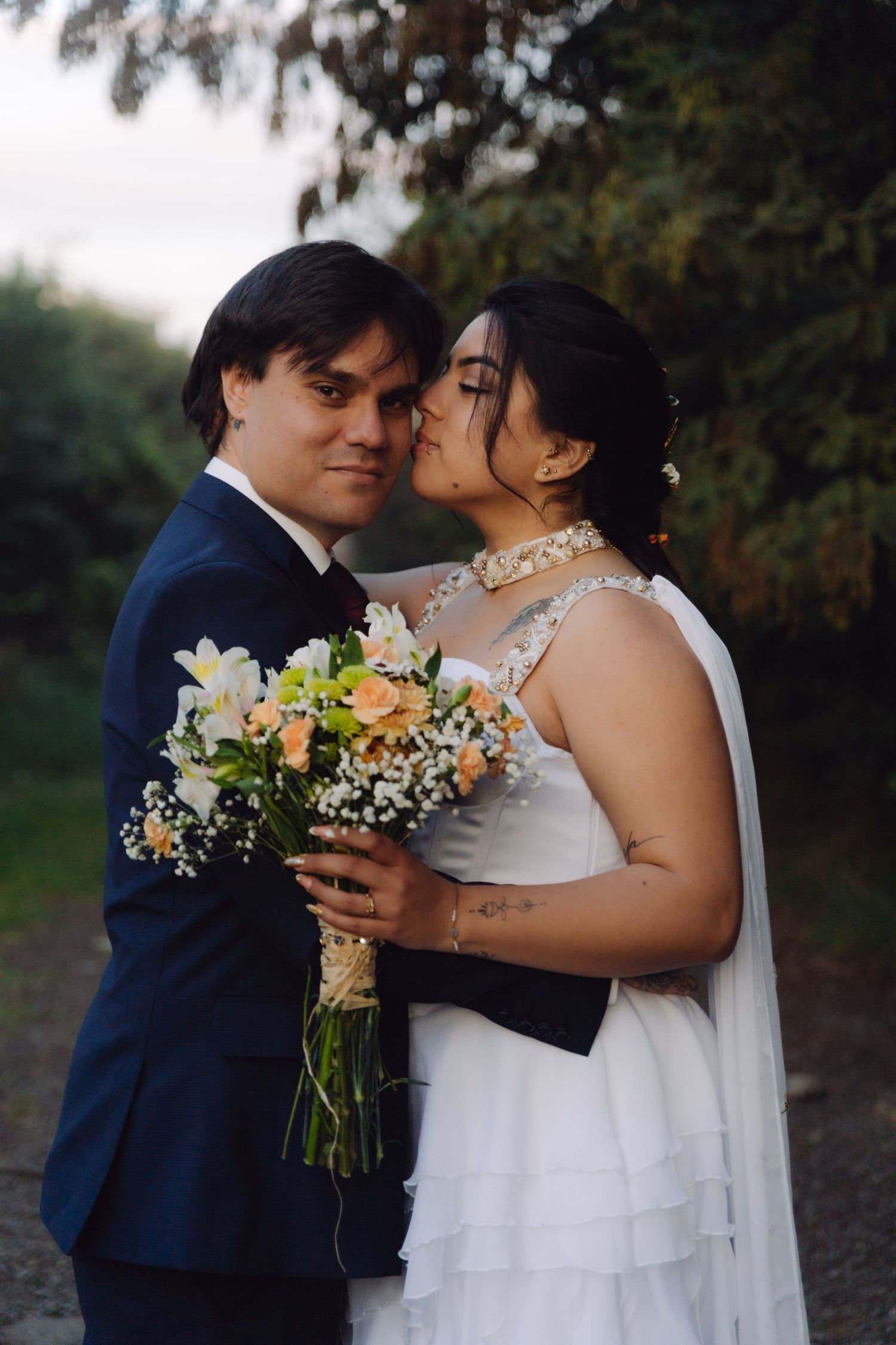 Bride kisses grooms cheek as they embrace outdoors holding a bouquet of peach and white flowers.