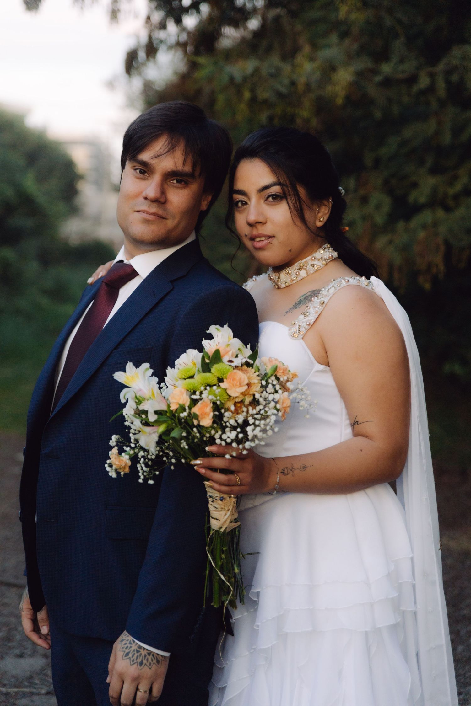 Bride and groom pose together outdoors, she holds a bouquet of peach and white blooms.