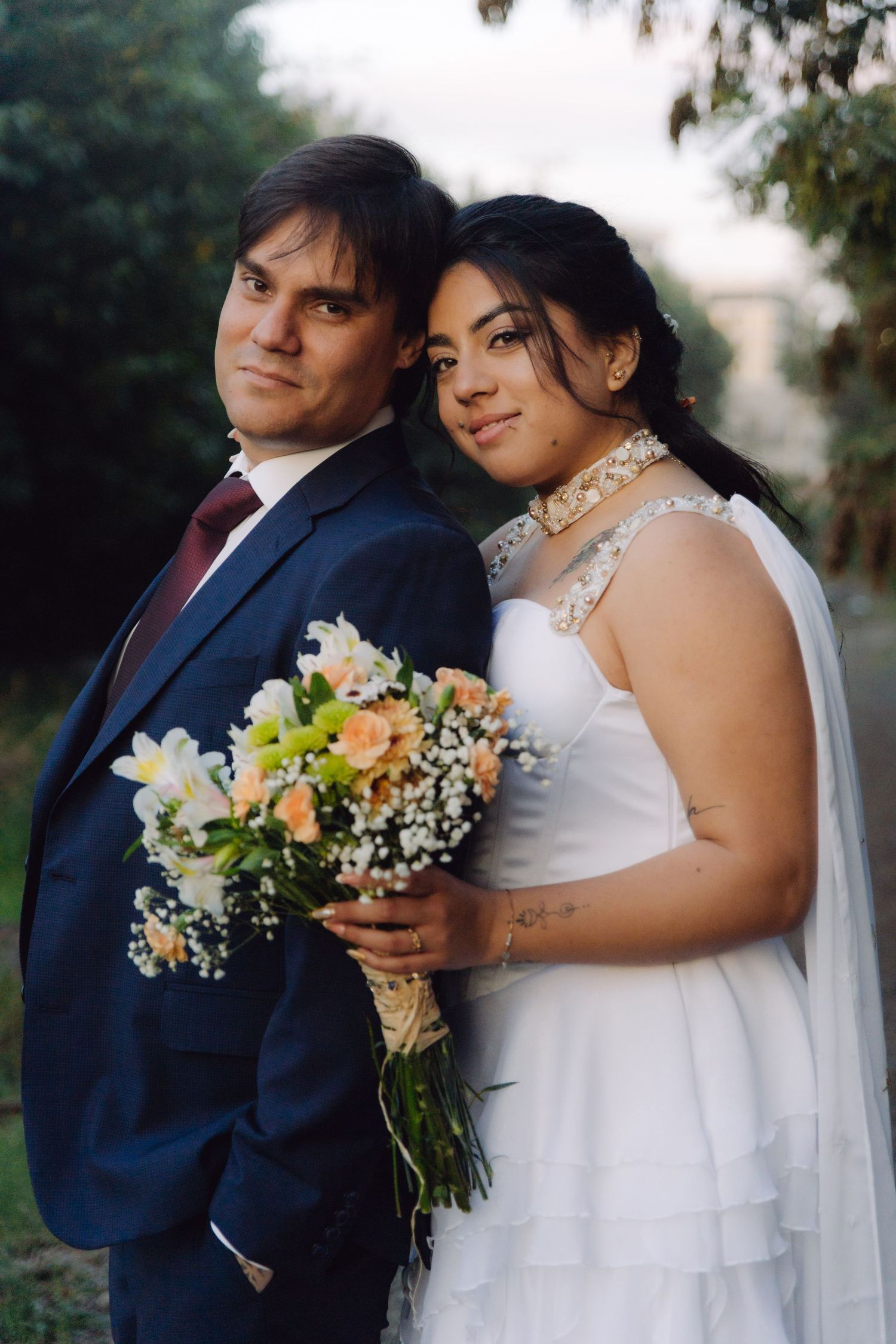 A bride leans against her groom as they pose together holding a floral bouquet at dusk.