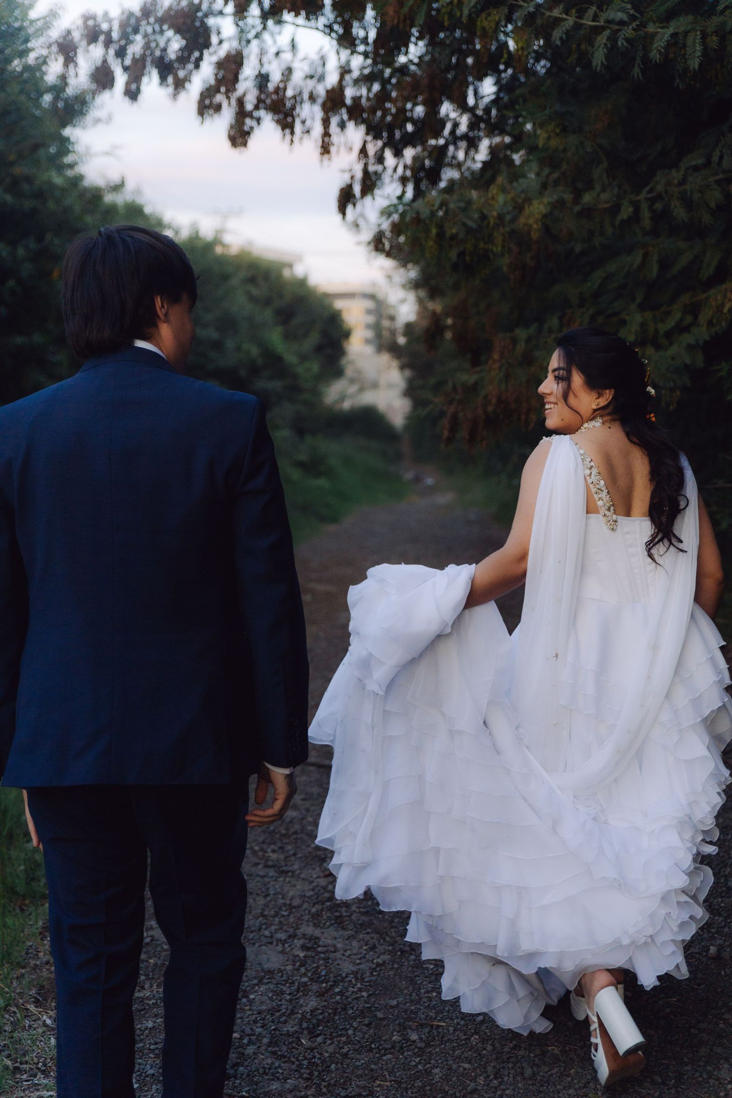 Bride smiles back at camera while walking with groom along a lush green tree-lined path.