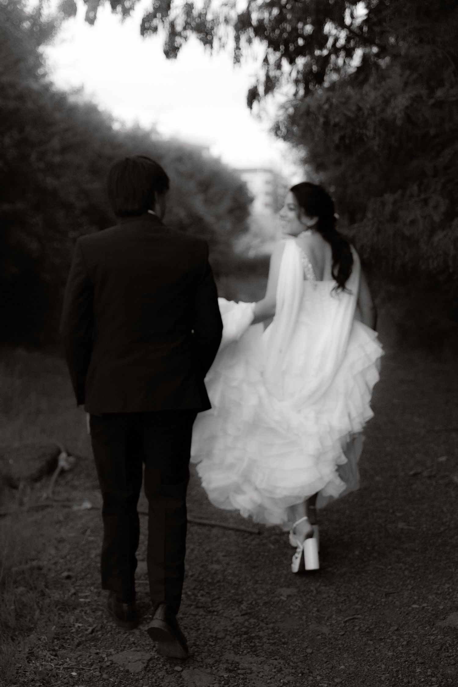 Black and white rear view of a bride and groom walking hand in hand along a tree-lined path.