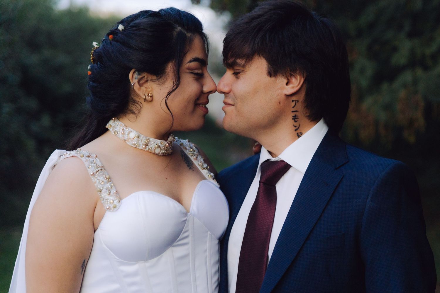 A bride and groom lean in close, nearly touching noses, smiling softly in an outdoor setting.