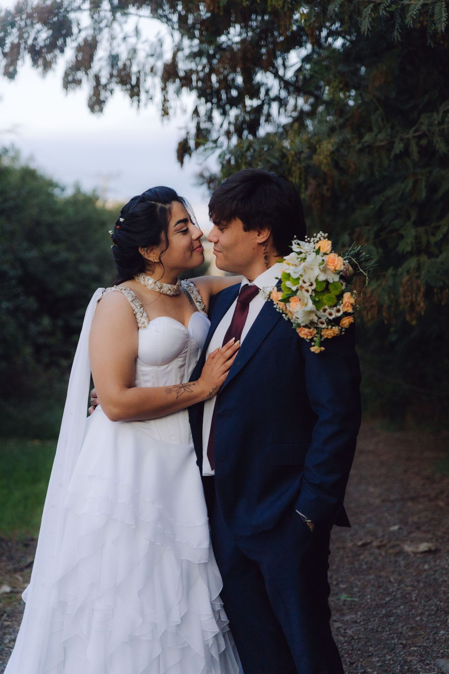 Romantic wedding couple leaning in for a kiss outdoors, groom holding bouquet of peach roses.