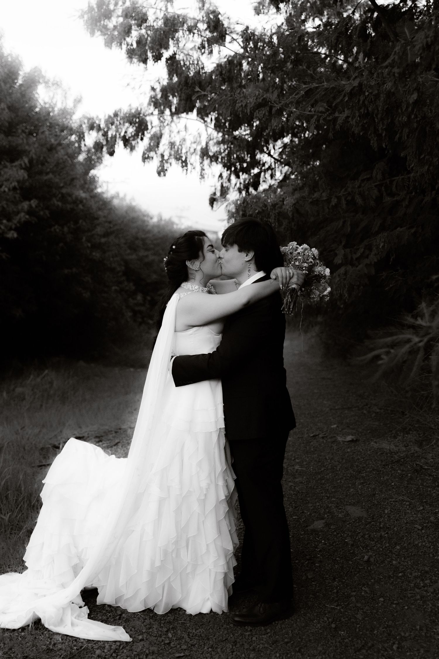 Black and white romantic wedding photo of couple kissing on a tree-lined path outdoors.