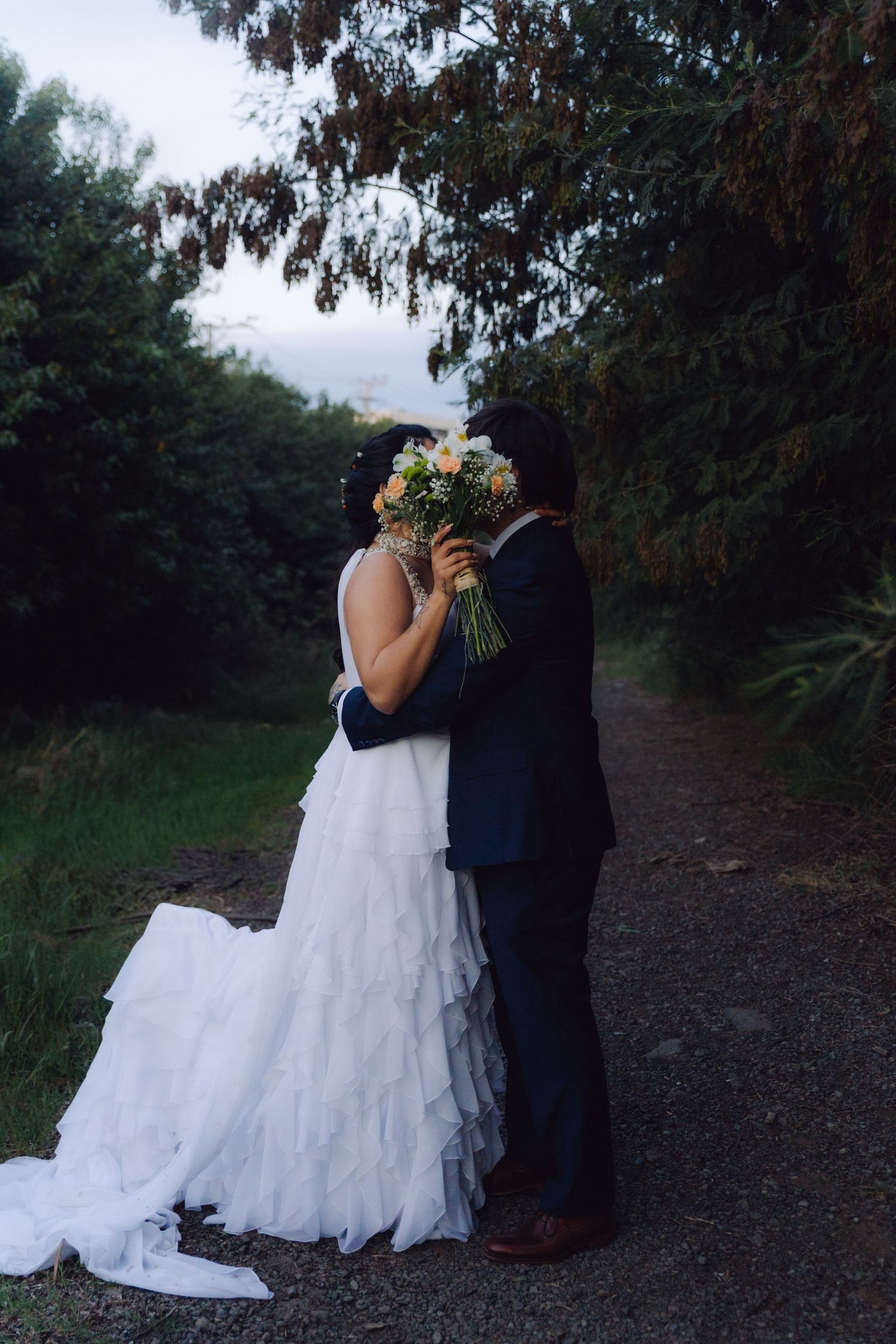 Bride and groom kissing behind a colorful bouquet on a scenic outdoor garden path.
