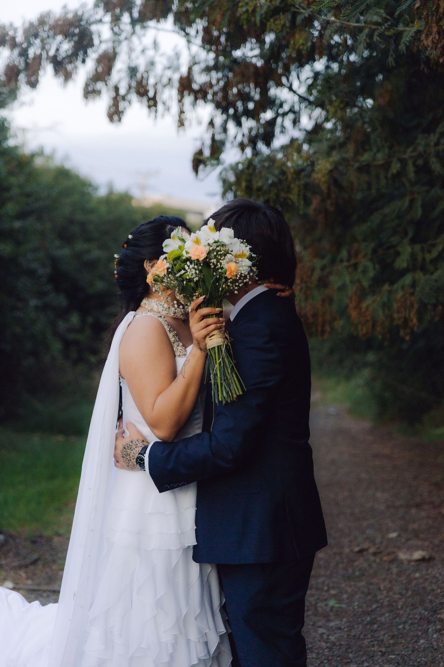 Wedding couple kissing behind a bouquet of peach and white flowers on a scenic outdoor path.