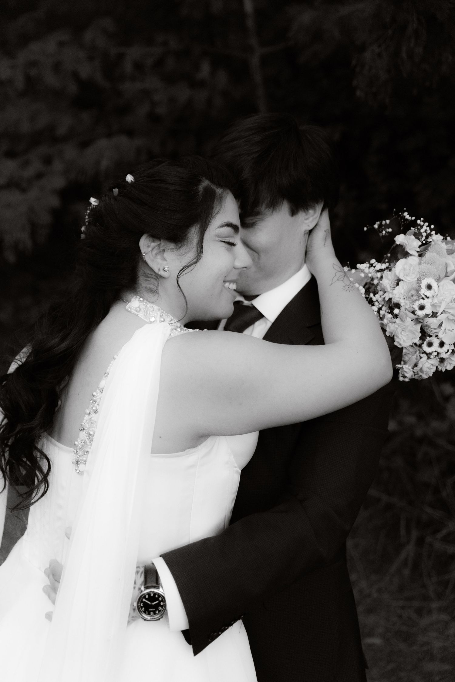 Black and white photo of smiling bride and groom touching foreheads, bride holding floral bouquet outdoors.