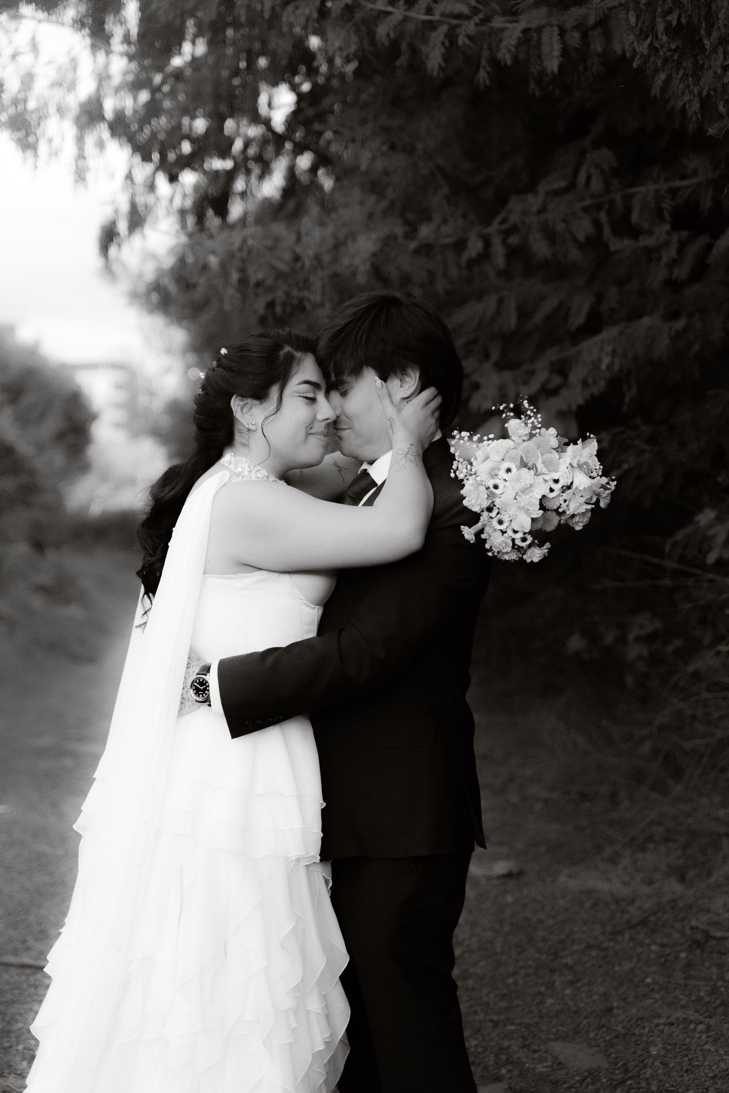 Black and white romantic wedding portrait of couple embracing and kissing outdoors among lush trees.