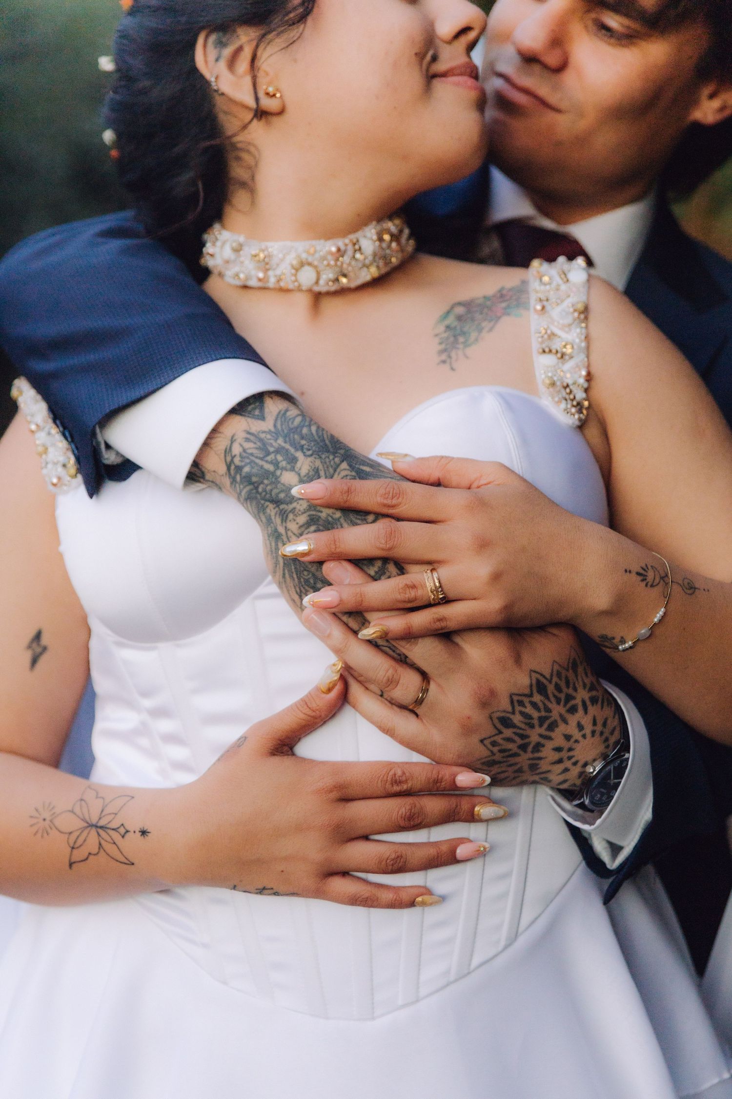 Close-up of tattooed bride and groom's hands with wedding rings, groom embracing bride in white dress.