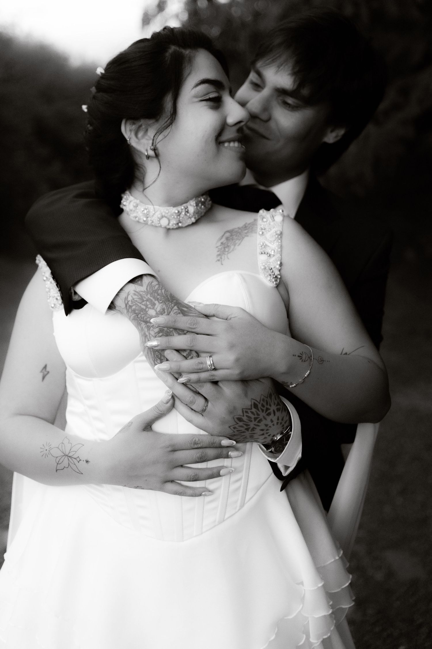Black and white photo of groom kissing tattooed bride's cheek while embracing her from behind outdoors.