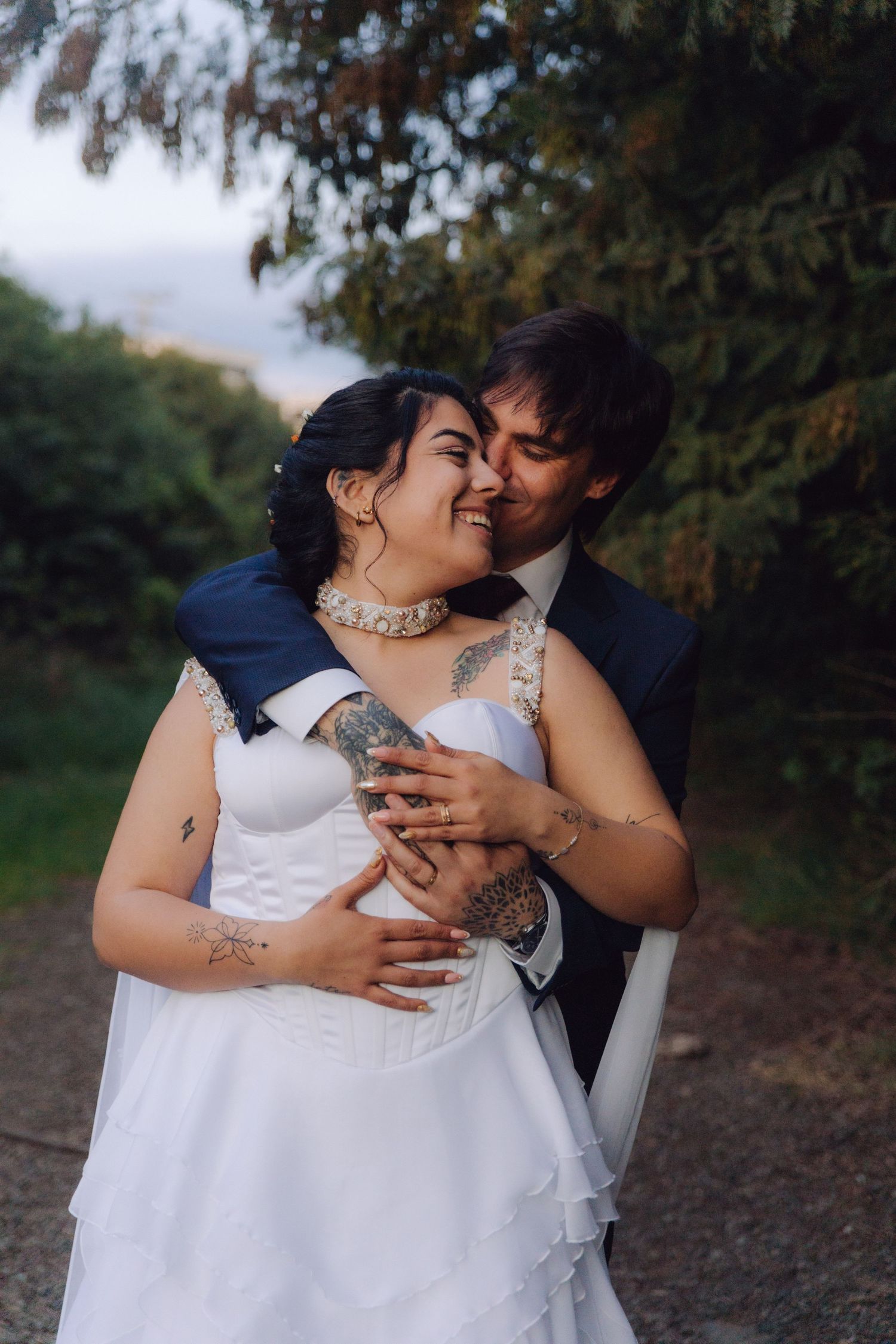 Groom kissing smiling tattooed bride on cheek while embracing her from behind in outdoor garden setting.