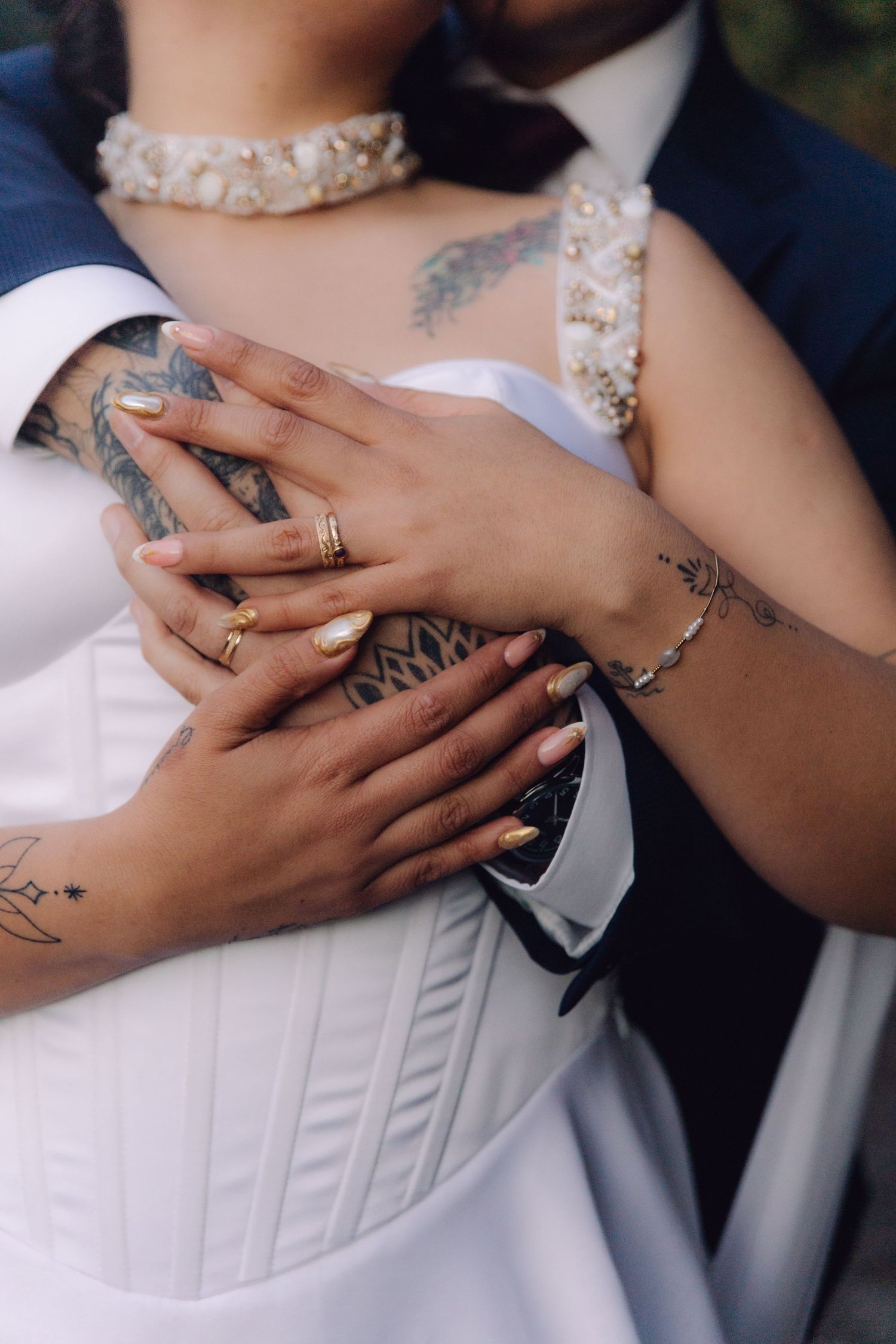 Close-up of tattooed newlyweds' hands with gold wedding rings, groom embracing bride in white corset dress.