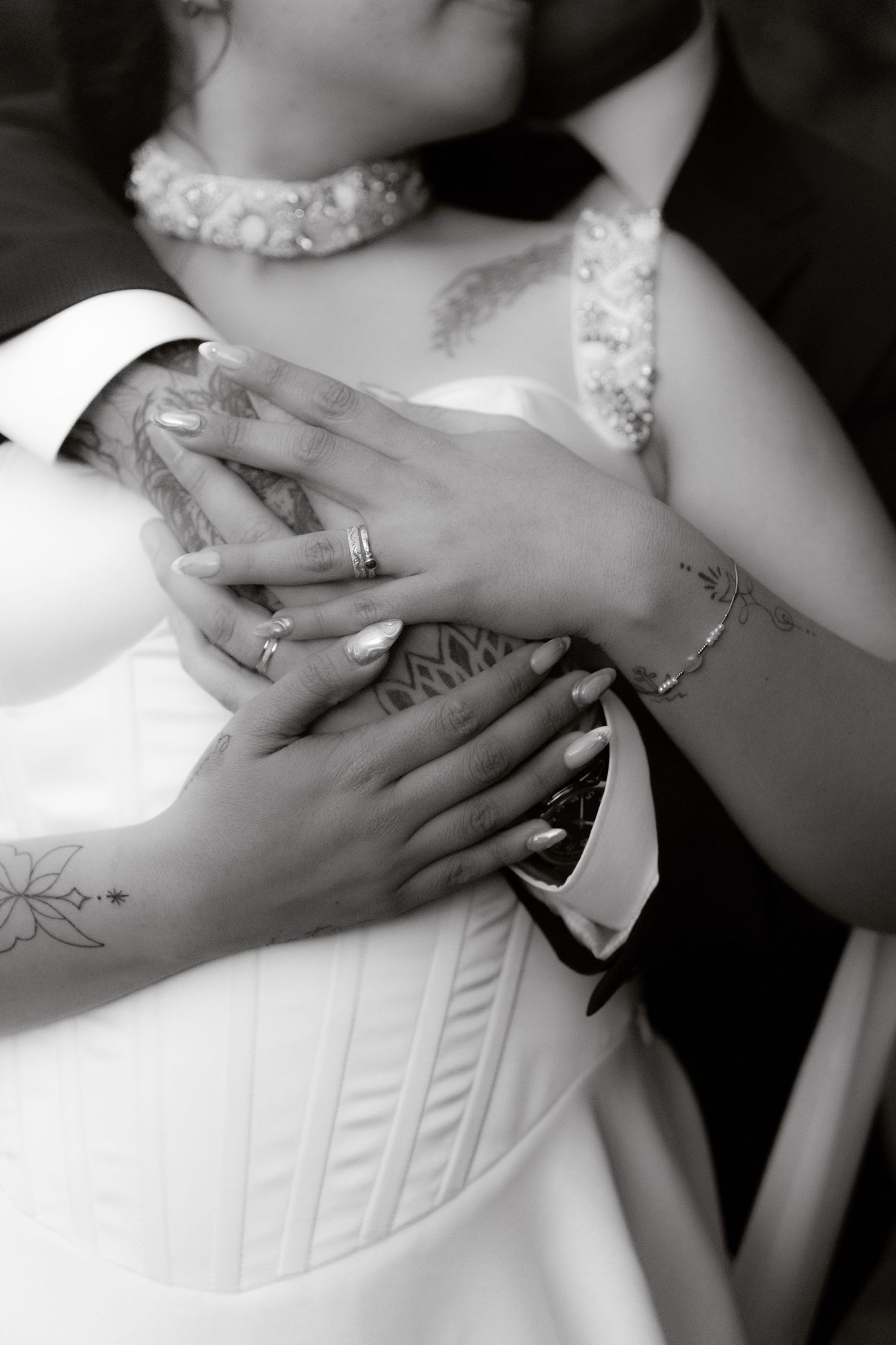 Black and white close-up of tattooed couple's hands with wedding rings, bride in white corset wedding dress.