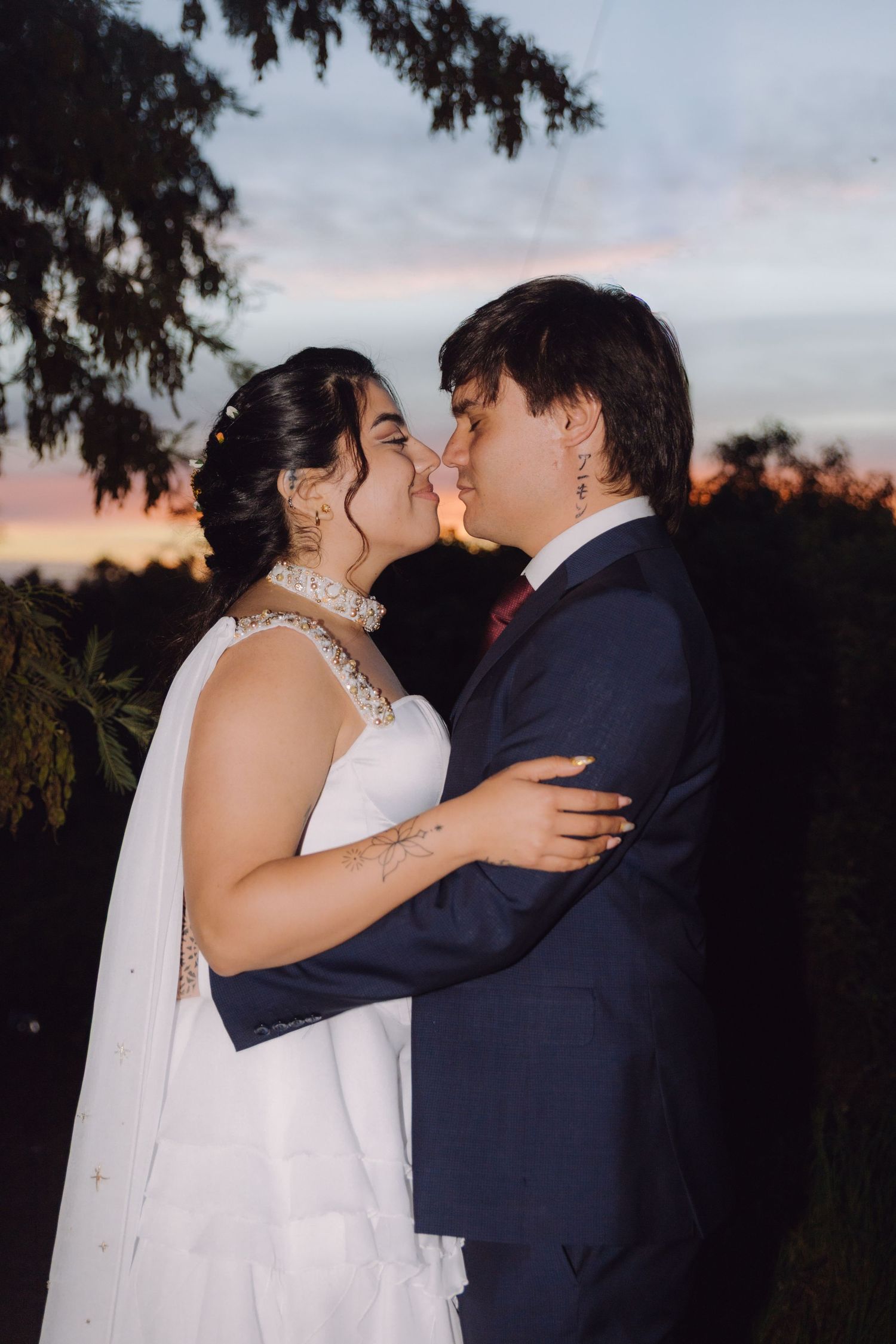 Bride and groom kissing at sunset with colorful sky backdrop during outdoor wedding photoshoot.