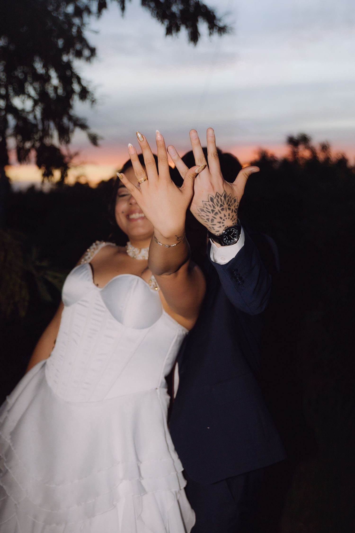 Newlyweds showing off wedding rings at sunset, bride in white gown and groom in navy suit.