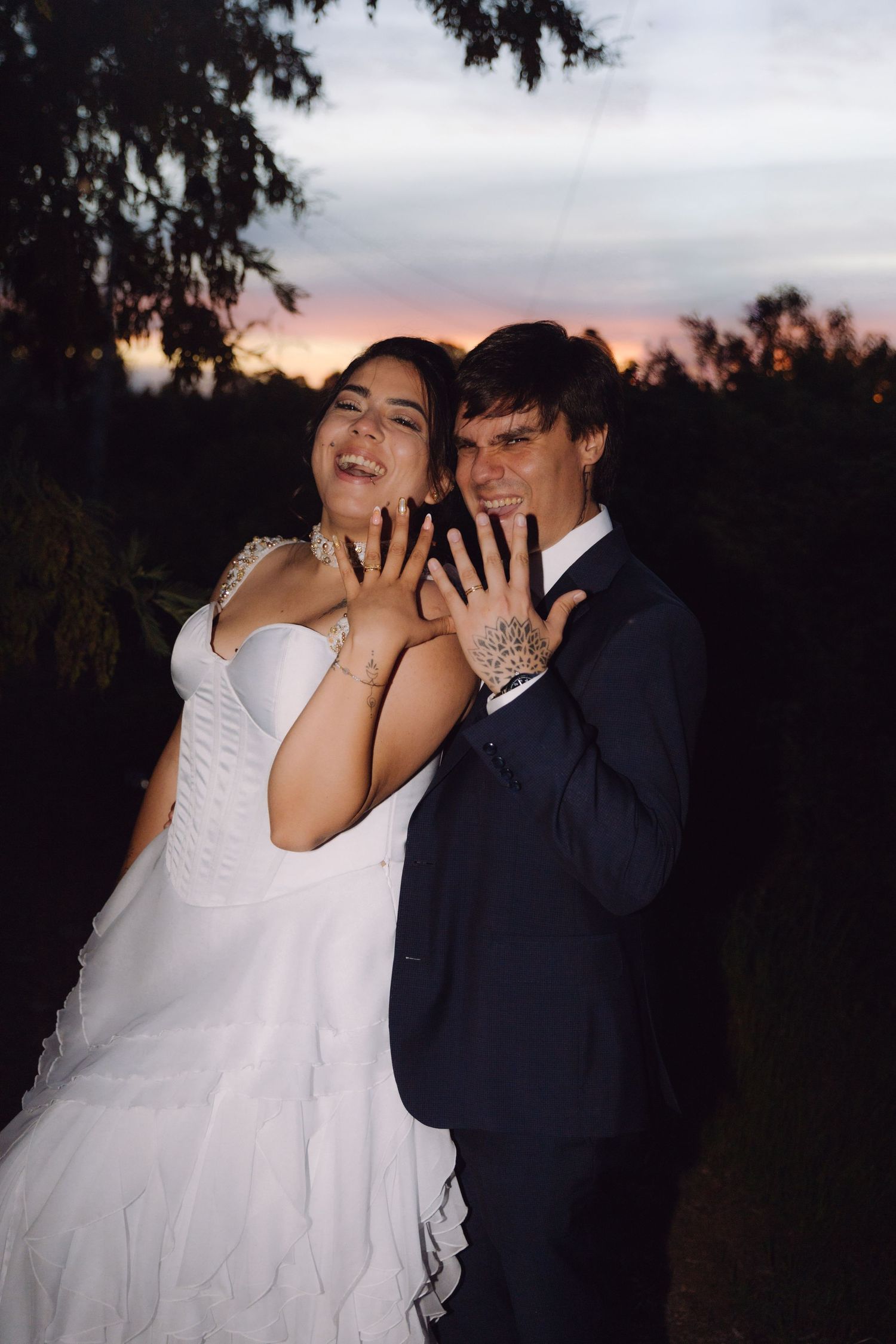 Happy bride and groom displaying wedding rings at dusk with colorful sunset sky behind them.