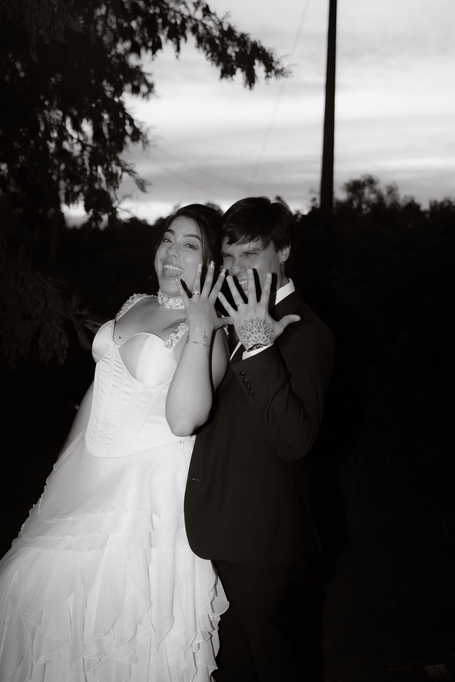 Black and white photo of smiling newlyweds showing wedding rings at twilight outdoors.