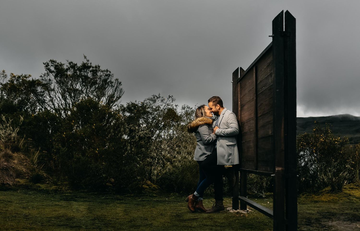 Couple sharing a romantic kiss beside a rustic wooden structure in a moody, overcast outdoor landscape.