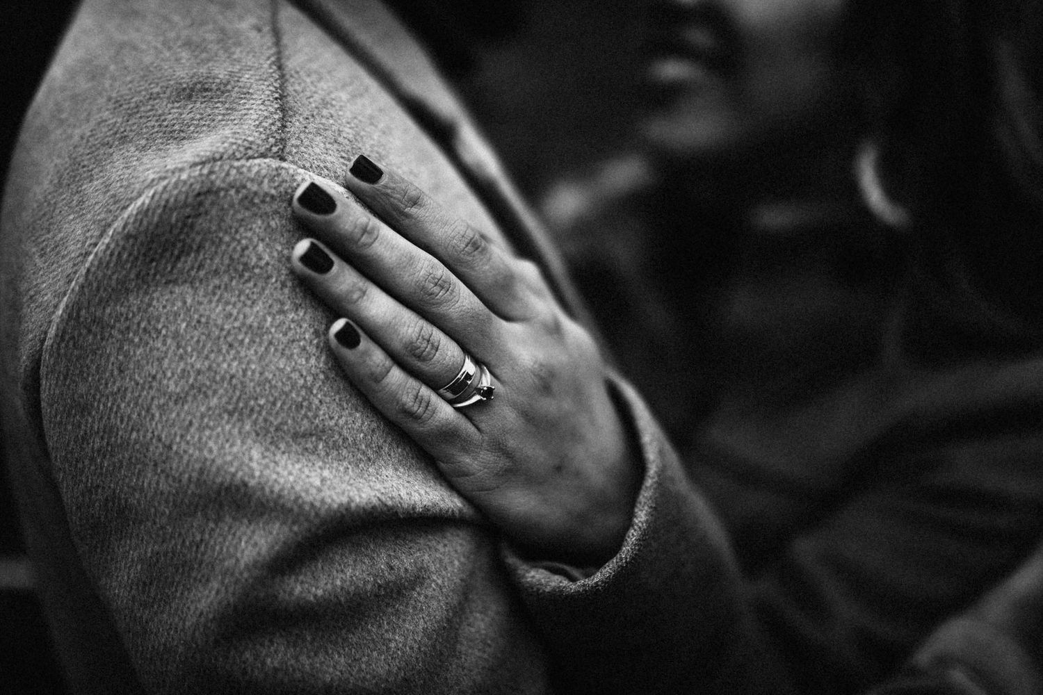 Black and white close-up of a hand with dark nail polish and a ring resting on a gray wool coat sleeve.
