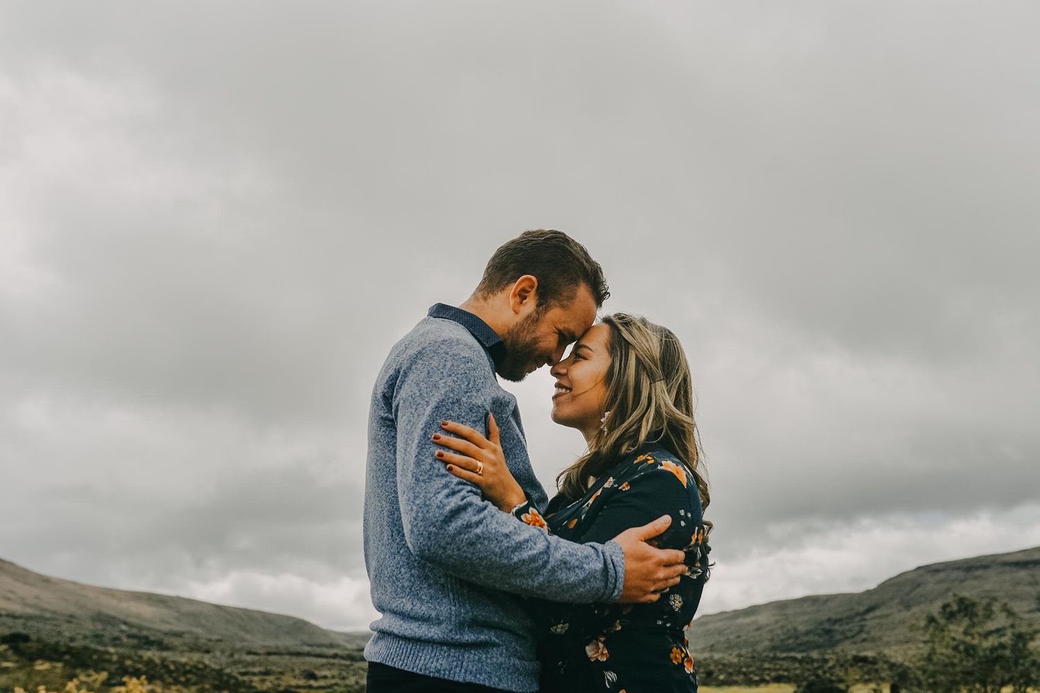 Romantic couple embracing outdoors under cloudy skies in a rugged landscape, touching foreheads in an intimate moment.