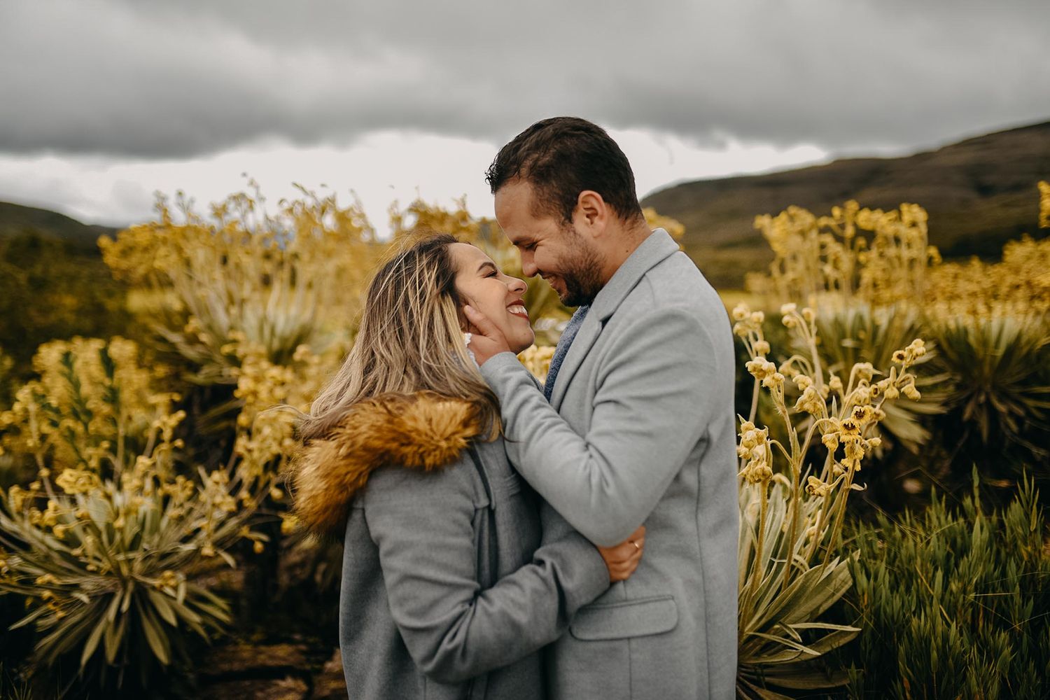 Couple in matching gray suits embracing outdoors among yellow wildflowers with misty mountains in the background.