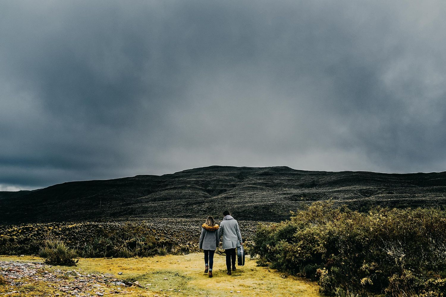 Two hikers walk hand-in-hand through a rugged highland landscape under dramatic stormy skies.