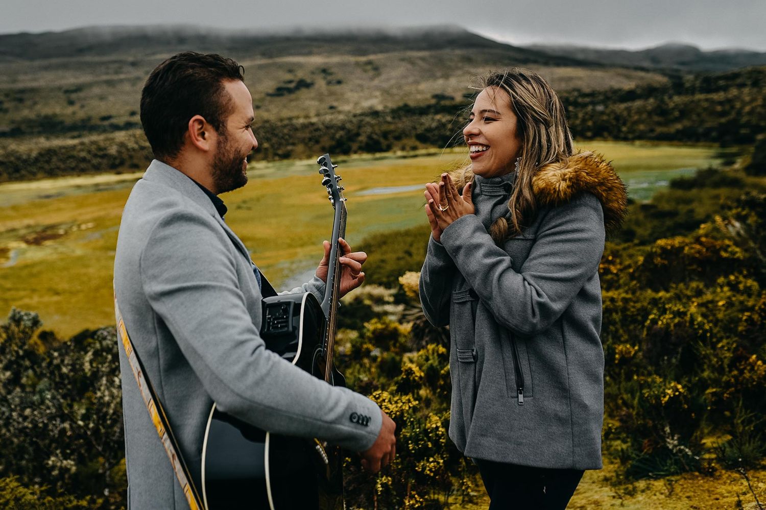 Man playing acoustic guitar for a smiling woman outdoors with scenic green mountain landscape in the background.