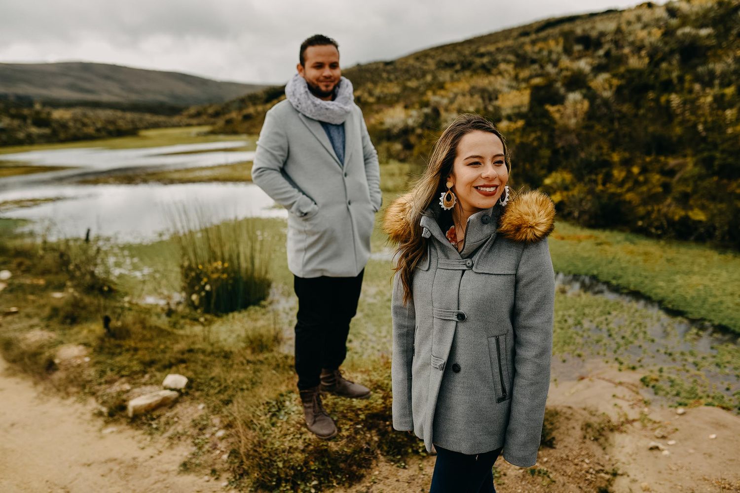 Couple in matching gray coats standing on a scenic mountain trail beside a tranquil lake on a cloudy day.