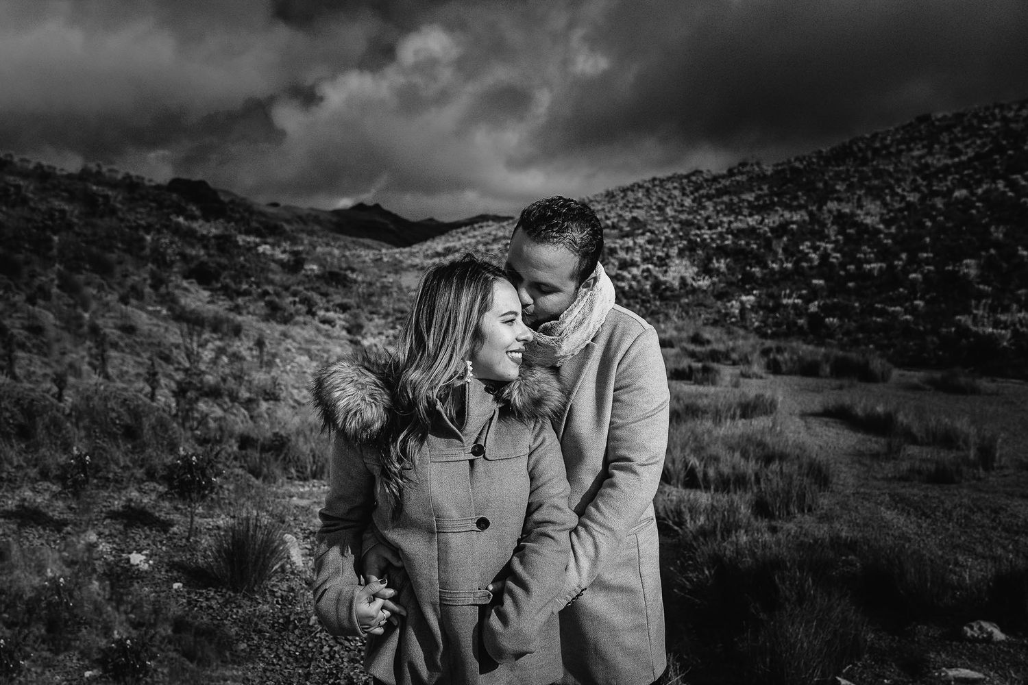 Black and white photo of a couple sharing a romantic moment in a dramatic highland landscape under stormy skies.