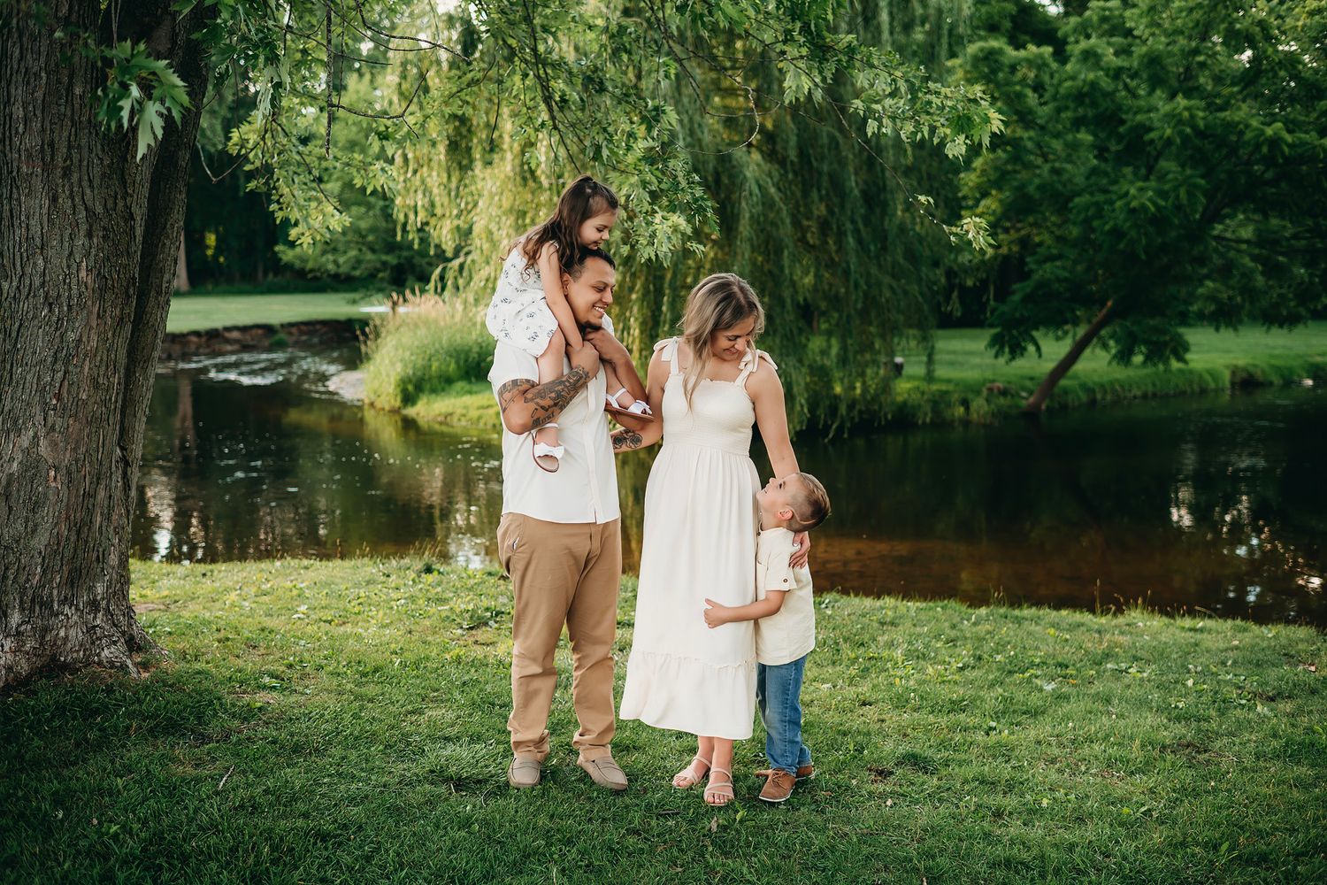 Family of four enjoying a sunny day in a lush green park near a pond, dressed in neutral tones.