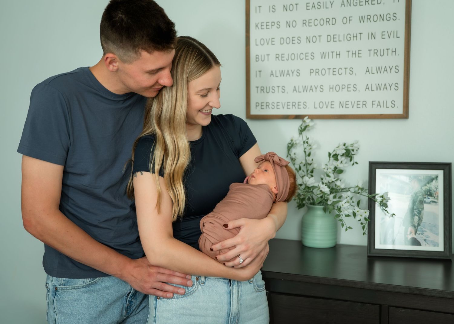 A couple smiles lovingly at their newborn baby wrapped in a pink swaddle, standing near a dresser with greenery.
