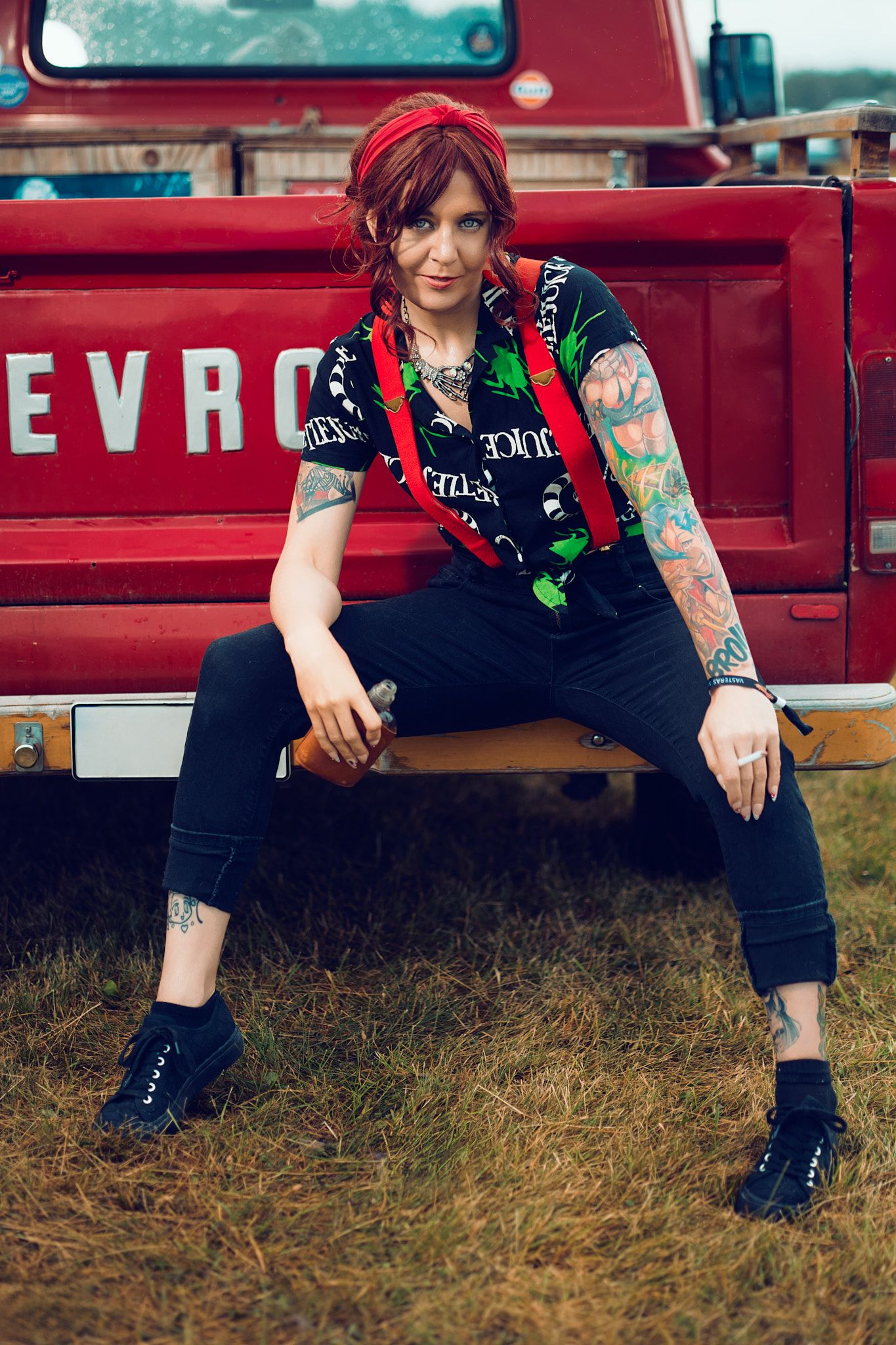 Tattooed woman with red hair in black outfit and suspenders sitting on vintage red Chevrolet truck bumper outdoors.