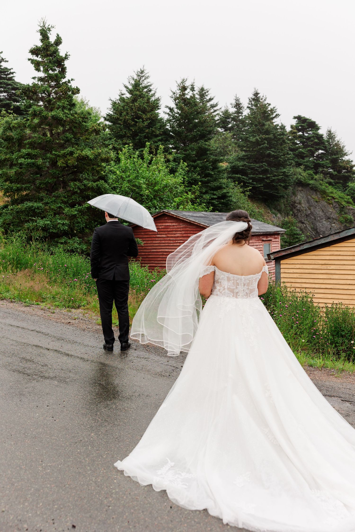 Bride in white gown and veil walks with groom holding umbrella on rainy wedding day outdoors.