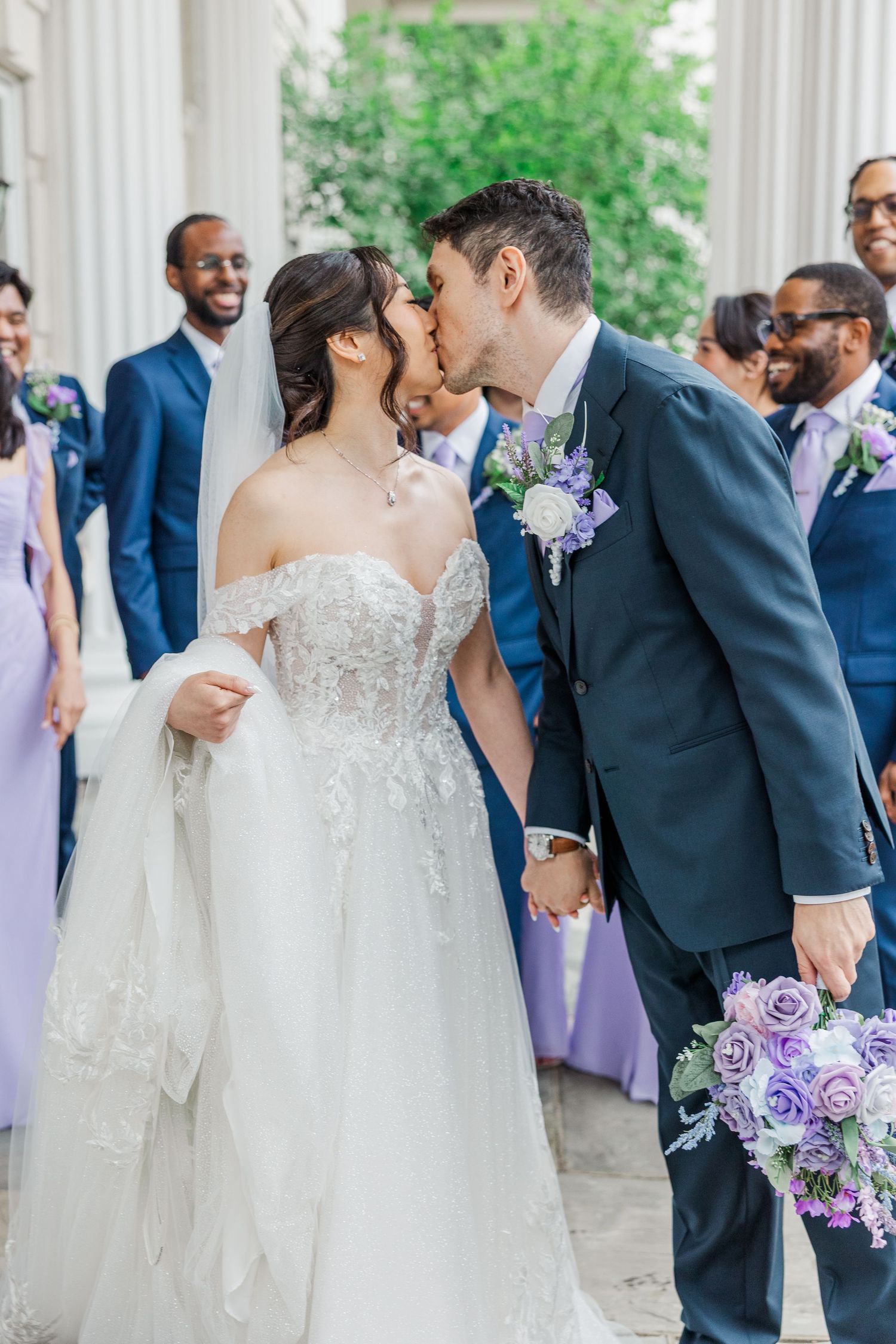 Bride and groom share a kiss at wedding ceremony with bridal party in blue and lavender attire in background.