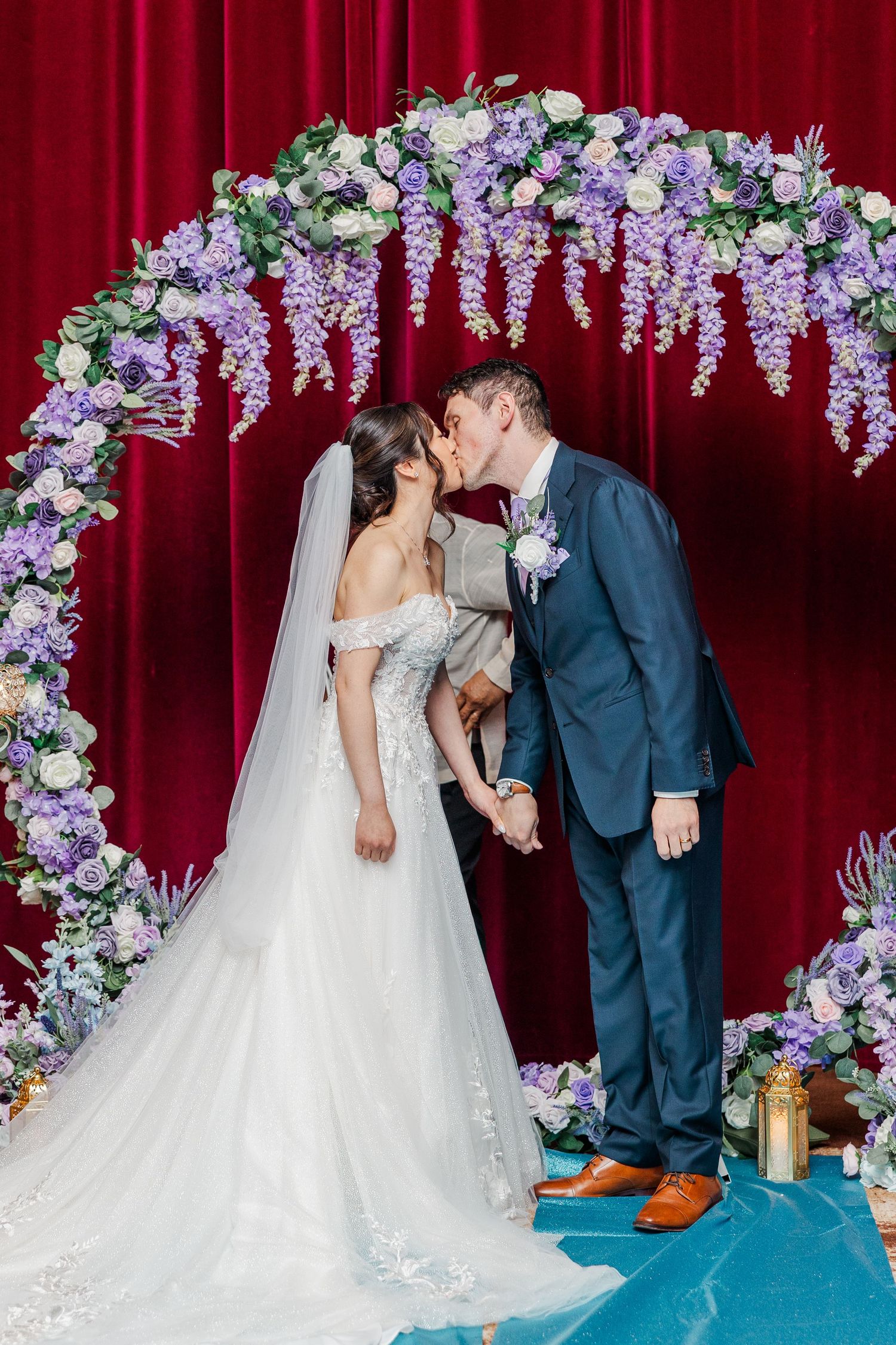 Bride and groom share first kiss under floral arch with purple flowers and red velvet backdrop at wedding ceremony.